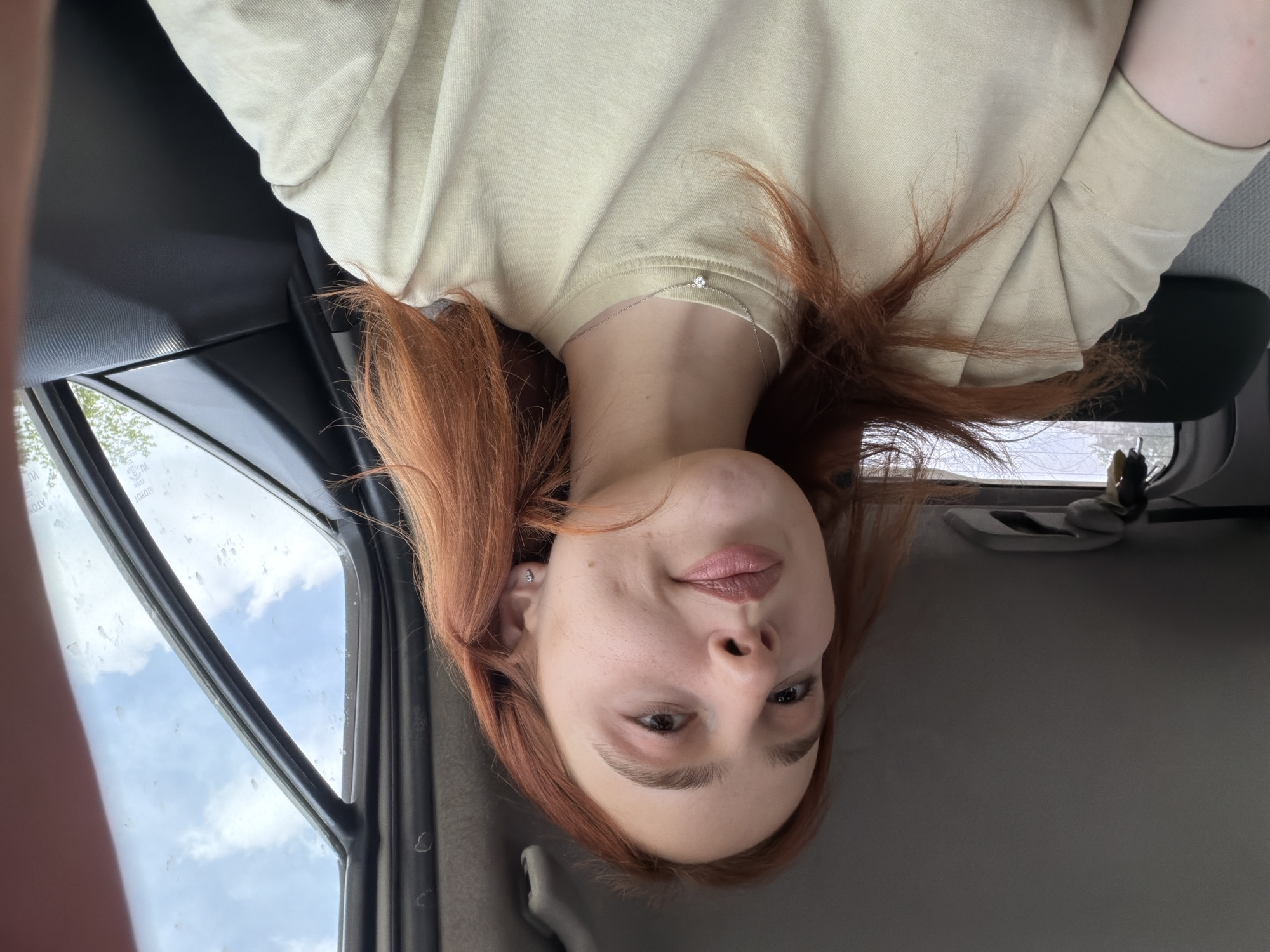 A young woman with red hair is in a car. She is wearing a light yellow t-shirt and a delicate necklace with a small pendant. The background shows a window with a view of the sky and a few trees. The scene may be within Tbilisi, Georgia, but there is insufficient information to determine the exact location within the city.