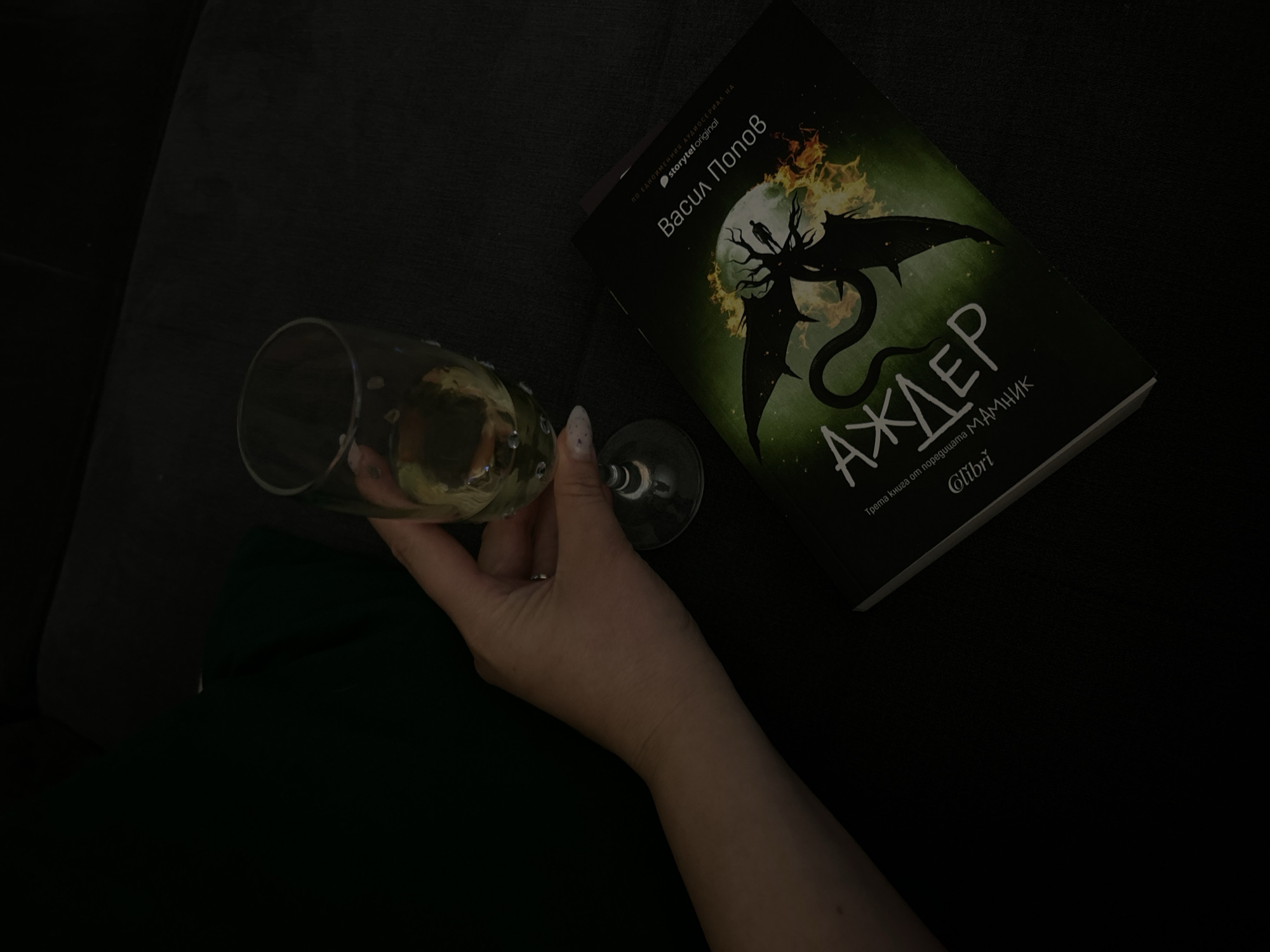 The image shows a hand holding a glass of champagne next to a book titled "Аждер", written by Васил Попов and published by Glibri. The book cover features a dark, mythical creature with fiery details, and the title is written in Cyrillic script. The setting appears to be indoors and dimly lit, with a dark surface, possibly a couch or table. The OCR indicates that the book is based on a storytel original audio series. No information about the specific location in Plovdiv, Bulgaria is available from the image.