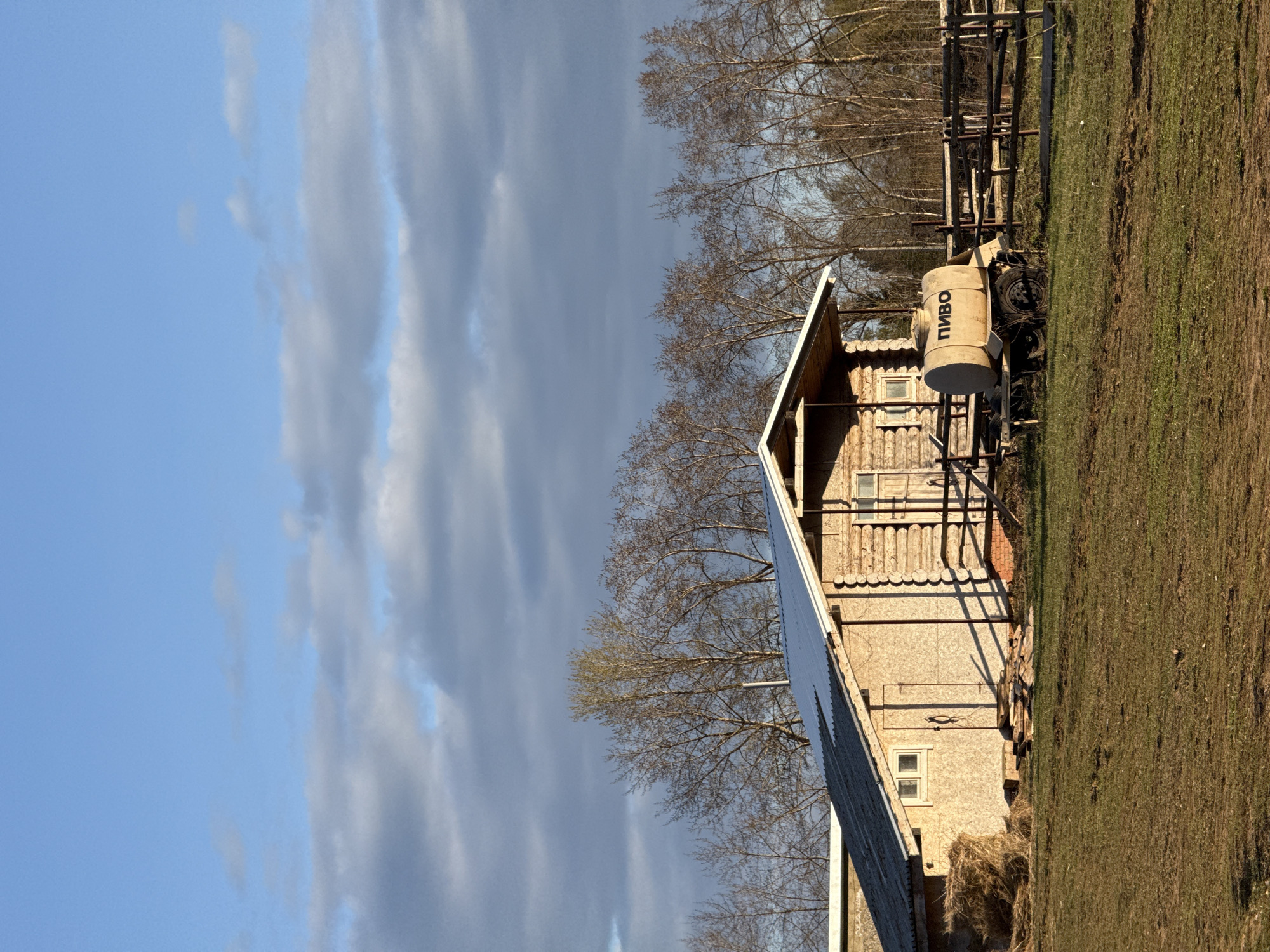 A daytime shot depicts a rural scene, likely in or near Galevo, Russia. A light-colored wooden building with a gabled roof dominates the right side of the frame. The building appears to be constructed from logs or planks, with visible windows and decorative elements along the eaves.
Adjacent to the building, a small tank truck with the Russian word "ПИВО" (Pivo, meaning beer) is parked on a grassy field. The field extends into the background, where a wooden fence and bare trees are visible.
The left side of the image features a cloudy sky, transitioning from blue to darker, overcast tones. The scene is bathed in sunlight, casting shadows and highlighting the textures of the building, grass, and trees. The overall impression is one of a simple, rustic setting, possibly a rural residential area or farm.