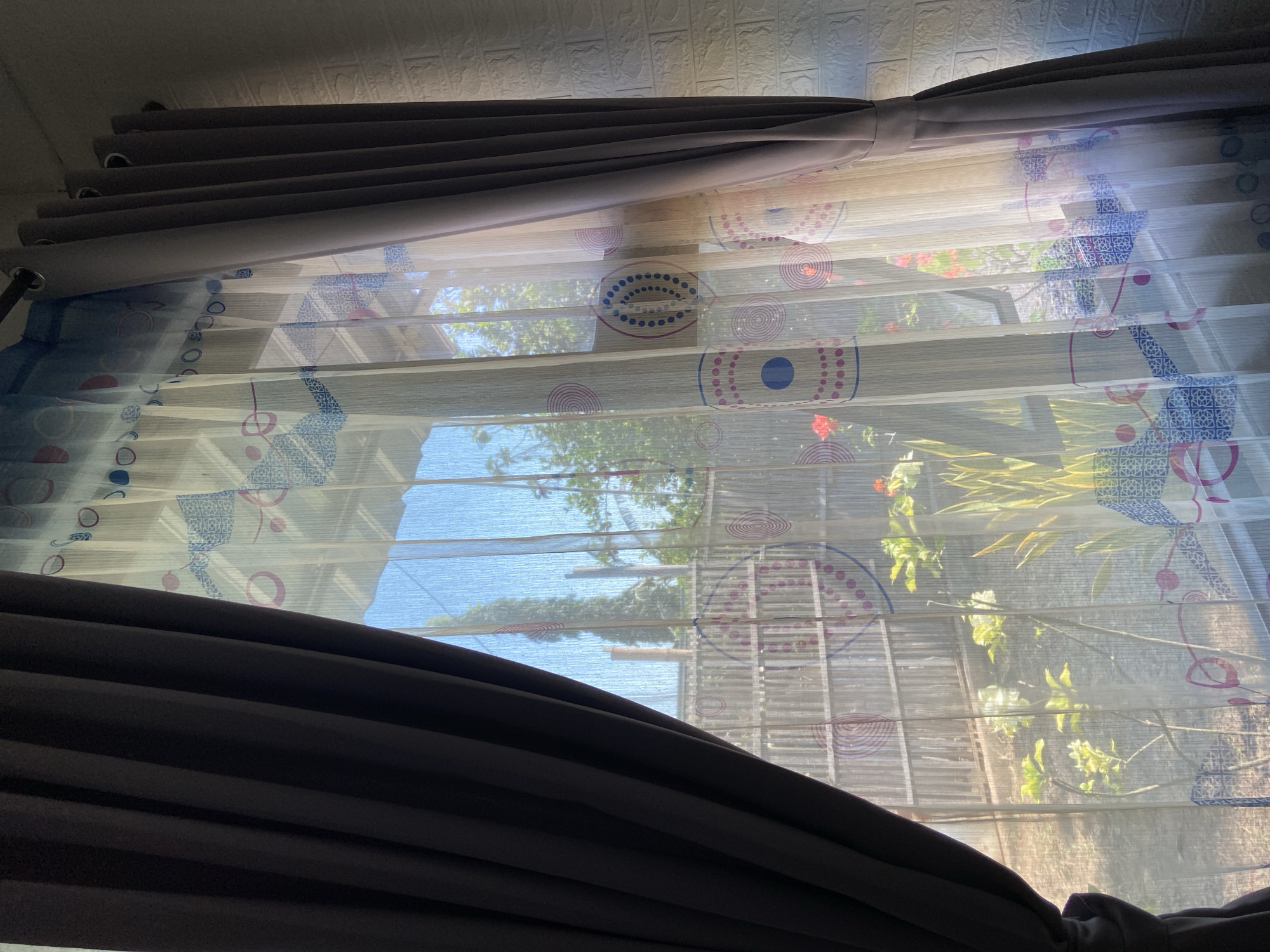 A window is covered by two sets of curtains, with sheer curtains featuring a repeating circular pattern in pink and blue layered in front of opaque, dark gray curtains. The sheer curtains allow sunlight to filter through, illuminating the room and the outside scene. Through the sheer curtains, a garden with green foliage and some red flowers is visible, along with a bamboo fence and trees. The bright sunlight casts shadows on the wall to the right of the window, which is covered in white brick-like wallpaper.