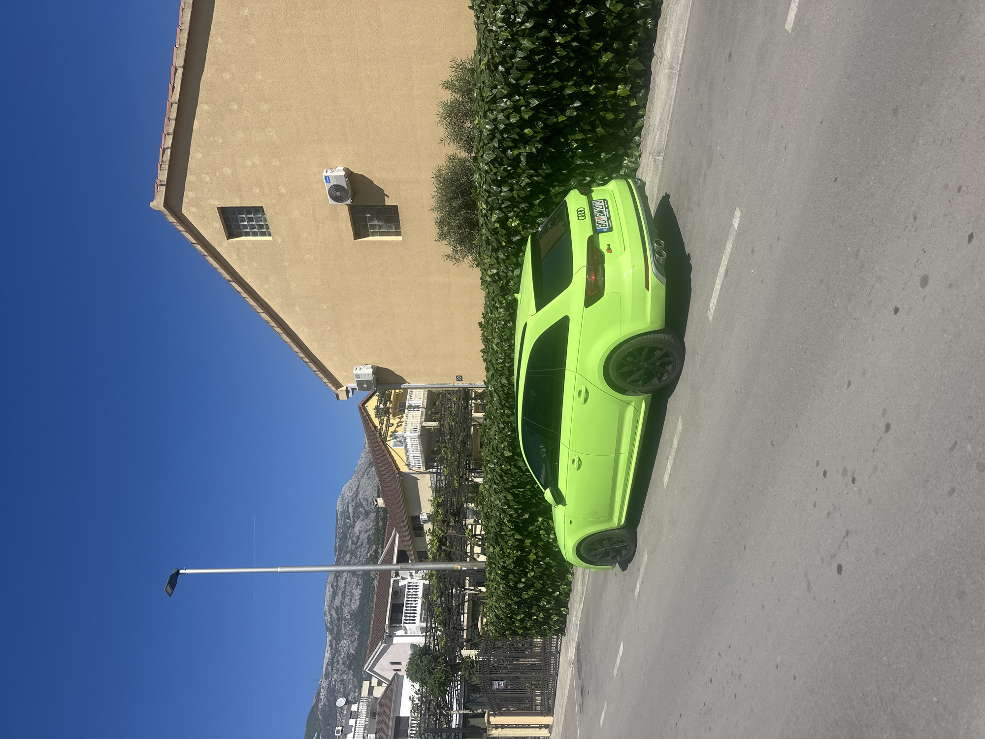 A brightly lit, landscape-oriented image captures a vivid lime green Audi car parked on the right side of a road, facing away from the viewer. The car appears to be an RS model, indicated by the "RS" badge on the rear. Its license plate, "BD•BW23", confirms the location as Budva, Montenegro. The car features black multi-spoke wheels and dual exhaust pipes.
To the left of the car, a light brown/tan building with a stucco-like finish stands under a clear, deep blue sky. This building has two square, gridded glass block windows and two external air conditioning units mounted on its wall. Green, dense foliage and bushes separate the road from the building.
Further into the background, a street light pole with a modern black fixture rises on the left. Beyond the immediate buildings and foliage, additional houses with white balconies and tiled roofs are visible, leading up to a range of rugged, rocky mountains under the clear sky. Strong shadows suggest bright sunshine, likely mid-day. The road itself is asphalt with faint white markings. A small sign with the number "20" is visible on a building further back. No people are present.