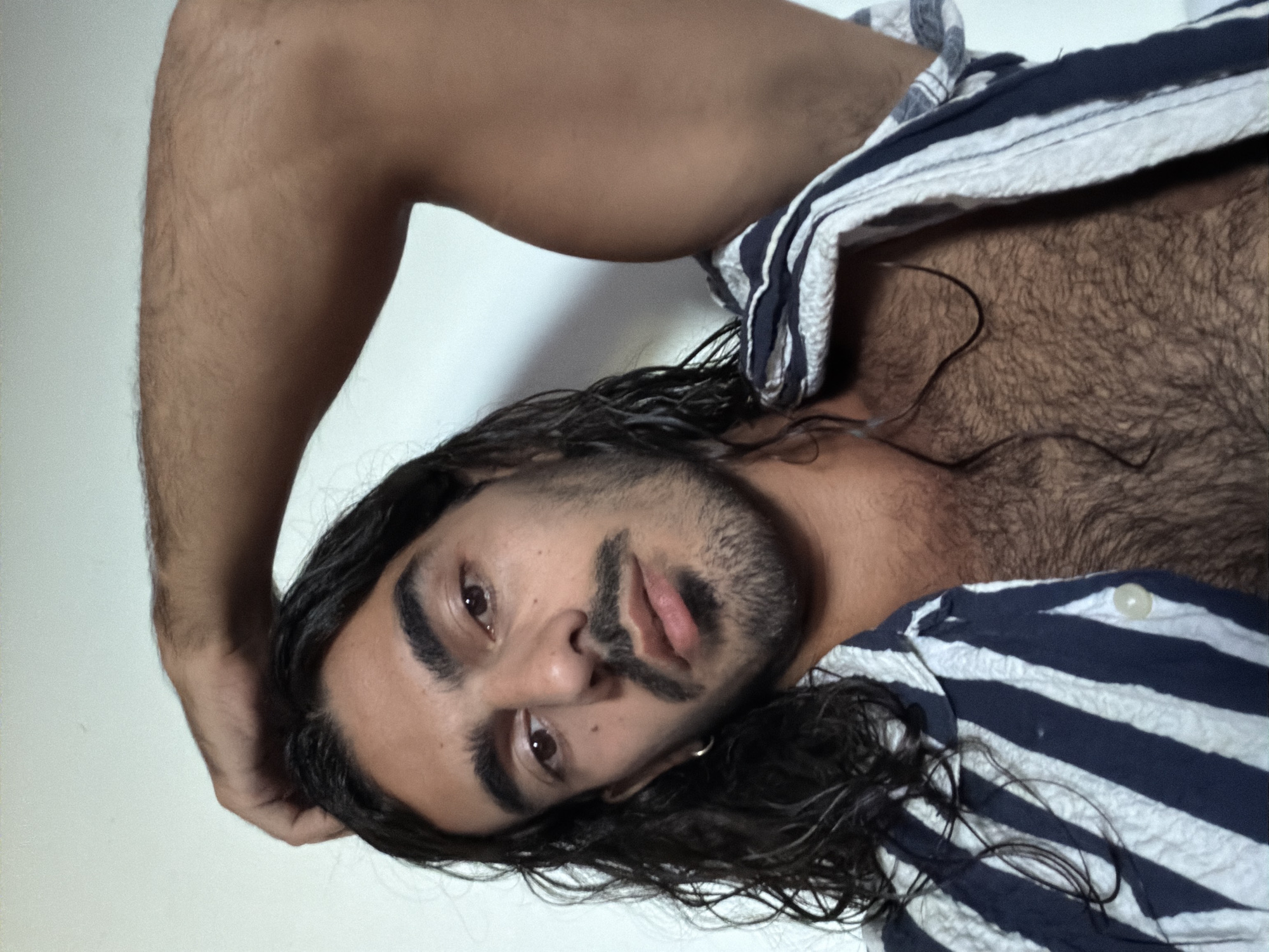 The image presents a close-up, waist-up portrait of a person, oriented on their side. The subject is an individual with long, dark, wavy hair that appears wet or damp, dark eyes, prominent dark eyebrows, a mustache, and a beard. Their gaze is directed intently at the viewer, with a neutral and somewhat thoughtful expression, and their lips are slightly parted. A small, silver hoop earring is visible in their right ear.
The person appears to be shirtless, revealing a chest and an arm covered in significant dark body hair. One arm is bent at the elbow, with the hand positioned near the back of the head. A blue and white striped shirt, possibly made of a textured fabric like seersucker, is partially draped over the person's torso, suggesting it is either off or loosely worn. One light-colored button is visible on the shirt near the shoulder area.
The background is a plain, light-colored surface, creating a minimalist indoor setting without any distinguishing features. The overall mood is one of relaxed introspection or direct engagement with the camera. There is no visible text in the image. The provided context indicates the location is North Kuta, Indonesia.