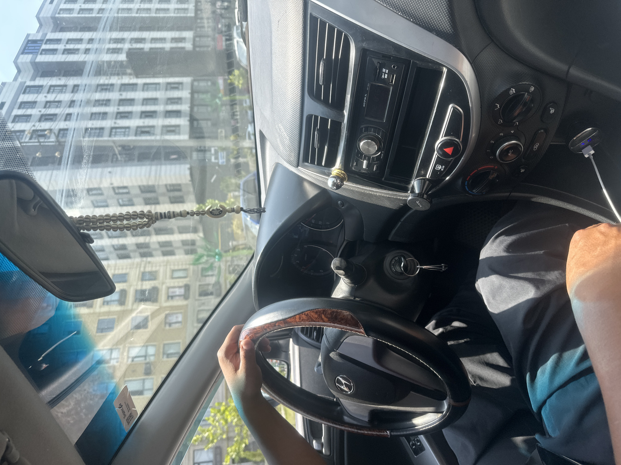 A first-person perspective shot from inside a car shows a person's hands on the steering wheel. The dashboard is visible, including the car's radio and air conditioning controls. A car mirror hangs from the ceiling, reflecting buildings in the background. Through the windshield, a street scene is visible, with tall buildings and trees. The image appears to be taken in a city, with elements suggesting a moderate urban environment. The location is identified as Manas, Kyrgyzstan.