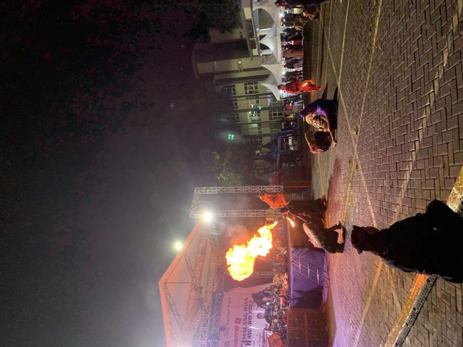 The scene is a nighttime outdoor performance in Kecamatan Batu, Indonesia, at the coordinates provided. A large flame shoots out from a fire-breathing performer standing on the stage, while several other performers wearing elaborate costumes and traditional masks are situated around the stage. The stage is made of metal and wood and is set up in front of a larger building with several windows. It is set up in front of the building. The performer is facing away from the camera, and the flame is directed toward the right side of the image. To the left of the stage, there are spectators seated on the ground. The performer is wearing a black costume with a red sash. The stage is decorated with a banner that reads "MALANG RAYA AWARD 2023". The image appears to have been taken during a nighttime performance.