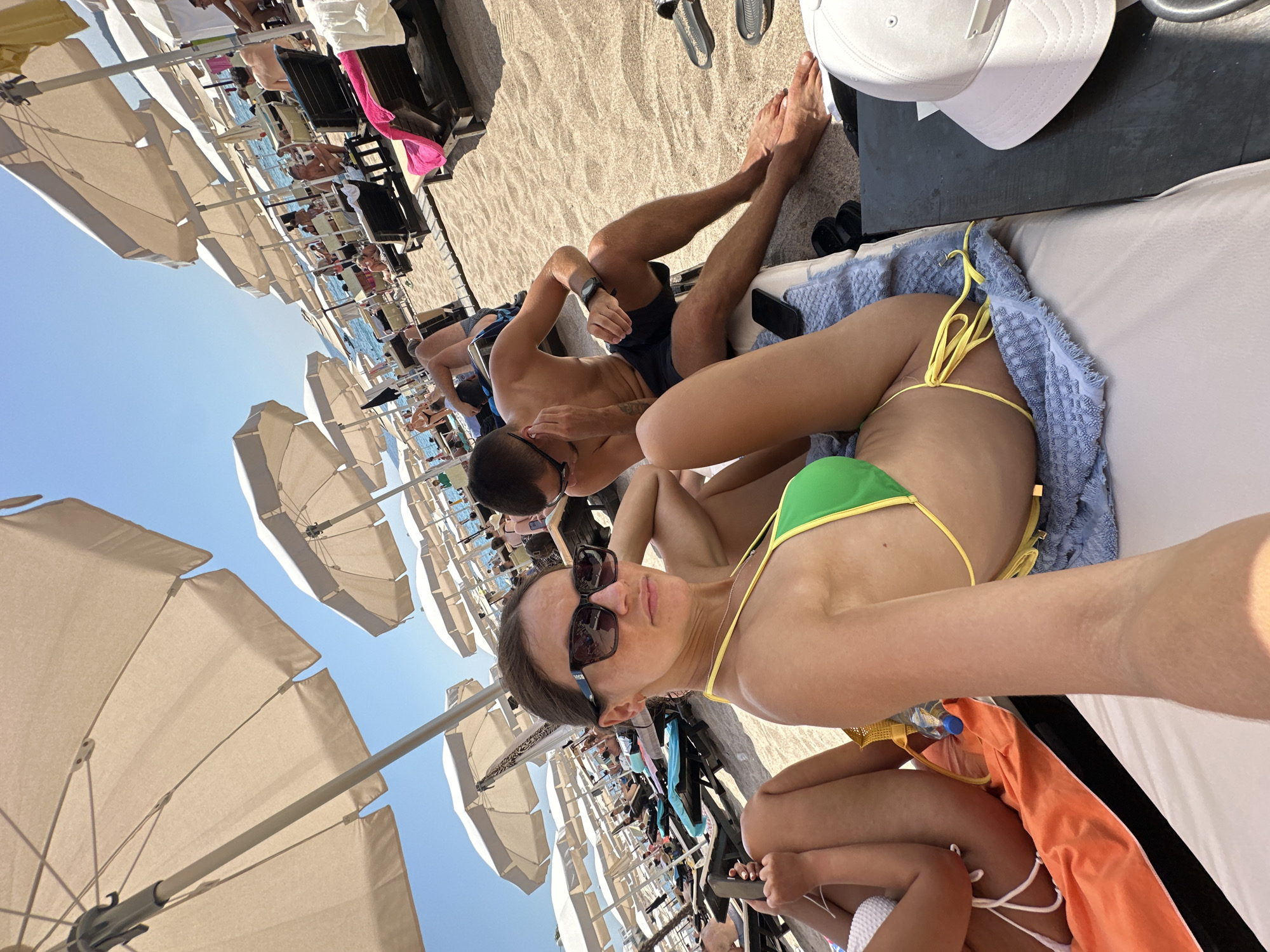 The image captures a sunny day on a crowded beach in Kamenovo, Montenegro. Several beach umbrellas provide shade over rows of lounge chairs occupied by people relaxing on the sand. In the foreground, a woman wearing sunglasses and a green bikini is seated on a lounge chair, looking towards the camera. Next to her, to the left, are the legs of another person in a white bikini. A man is seated on a lounge chair behind the woman in the green bikini, looking to his right. Various beach accessories such as towels, a water bottle, and a white cap are visible. The environment is a typical beach scene with sand, lounge chairs, and umbrellas under a clear blue sky.