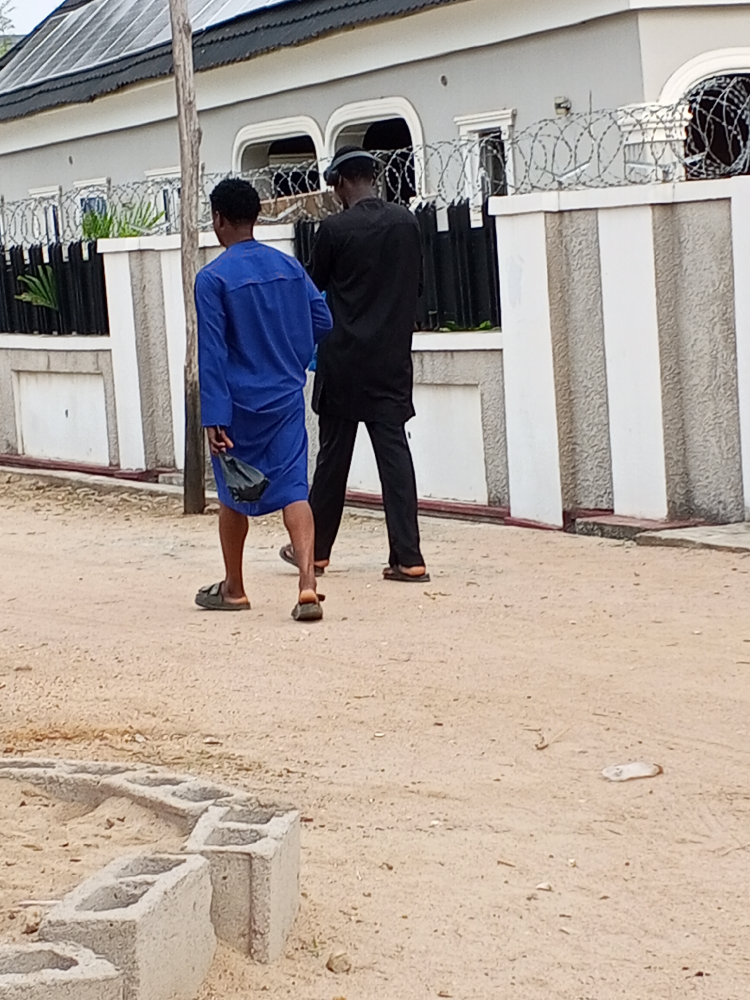 Two men are walking on a sandy road in Adonkolo, Nigeria, in front of a house with a fence topped with barbed wire. The man on the left is wearing a blue robe and sandals, and carrying a black plastic bag. The man on the right is wearing a black suit and sandals. In the foreground are a few concrete blocks.