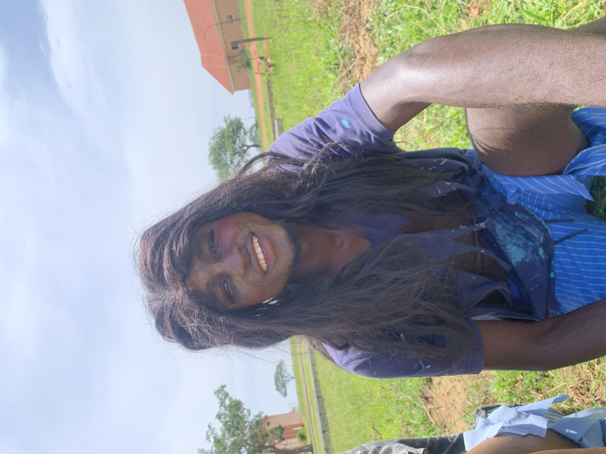 A young Black male is sitting outdoors in a grassy area, possibly in the city of Gwafan, Nigeria. He is wearing a torn, dark blue t-shirt with blue and white striped shorts. His face is painted with dark makeup, and he has a long, dark wig on. He is smiling broadly, showing his teeth. The background consists of green grass, some trees, and buildings in the distance under a bright, partly cloudy sky. The overall impression is that of a candid or posed photograph, possibly related to a cultural event or costume.