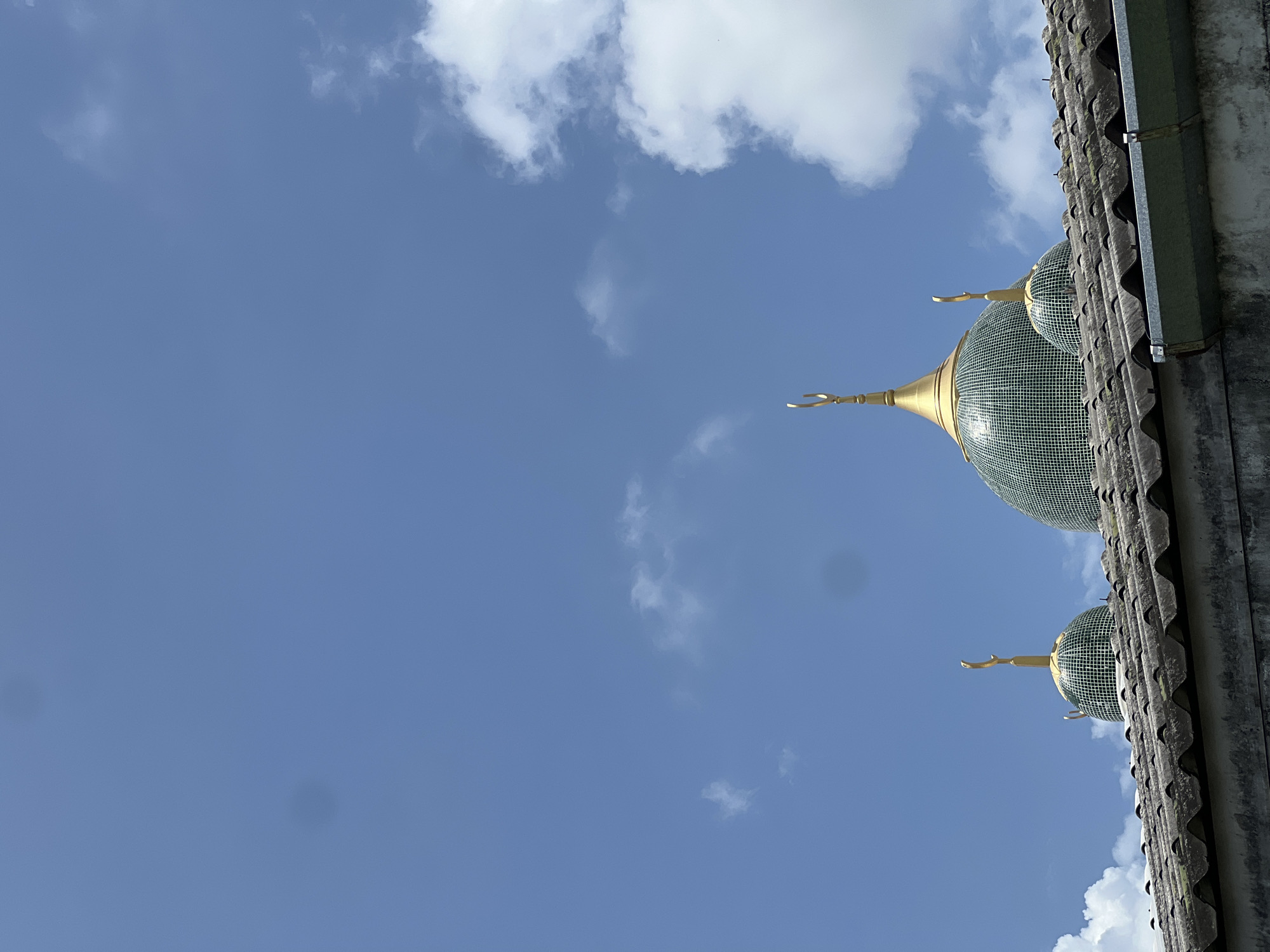 The image shows the tops of three green domes with golden crescent moons on top, likely part of a mosque, seen from a low angle against a blue sky with scattered white clouds. The domes are tiled with small green squares and have a mosaic-like appearance. They rise above a weathered, grey tiled roof. The scene is likely in the city of Choeng Thale, Thailand, given the architectural style and commonality of mosques in the region. There are no people visible in the image. The overall impression is of a sunny day with clear skies.
