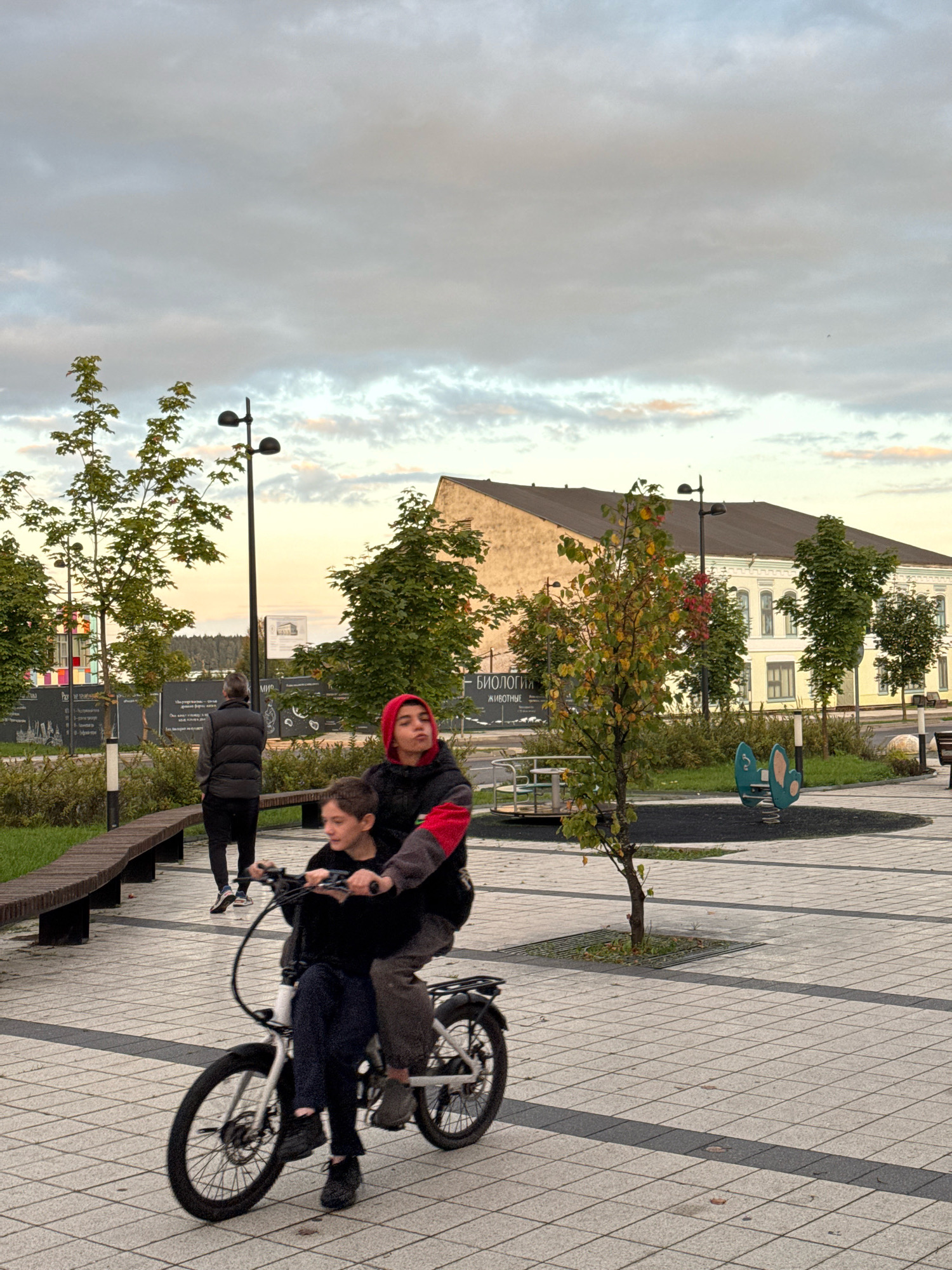 Two young people ride an electric bicycle on a paved pathway in Valday, Russia. The person in front, wearing dark clothing, is pedaling. The person behind, wearing a red hooded sweatshirt with a black body and red sleeves, is seated on the handlebars. They are in a public park with trees, benches, a lamppost, and playground equipment in the background. A building with a yellow facade and a dark roof is visible behind the trees. The sky is overcast with hints of sunset colors.