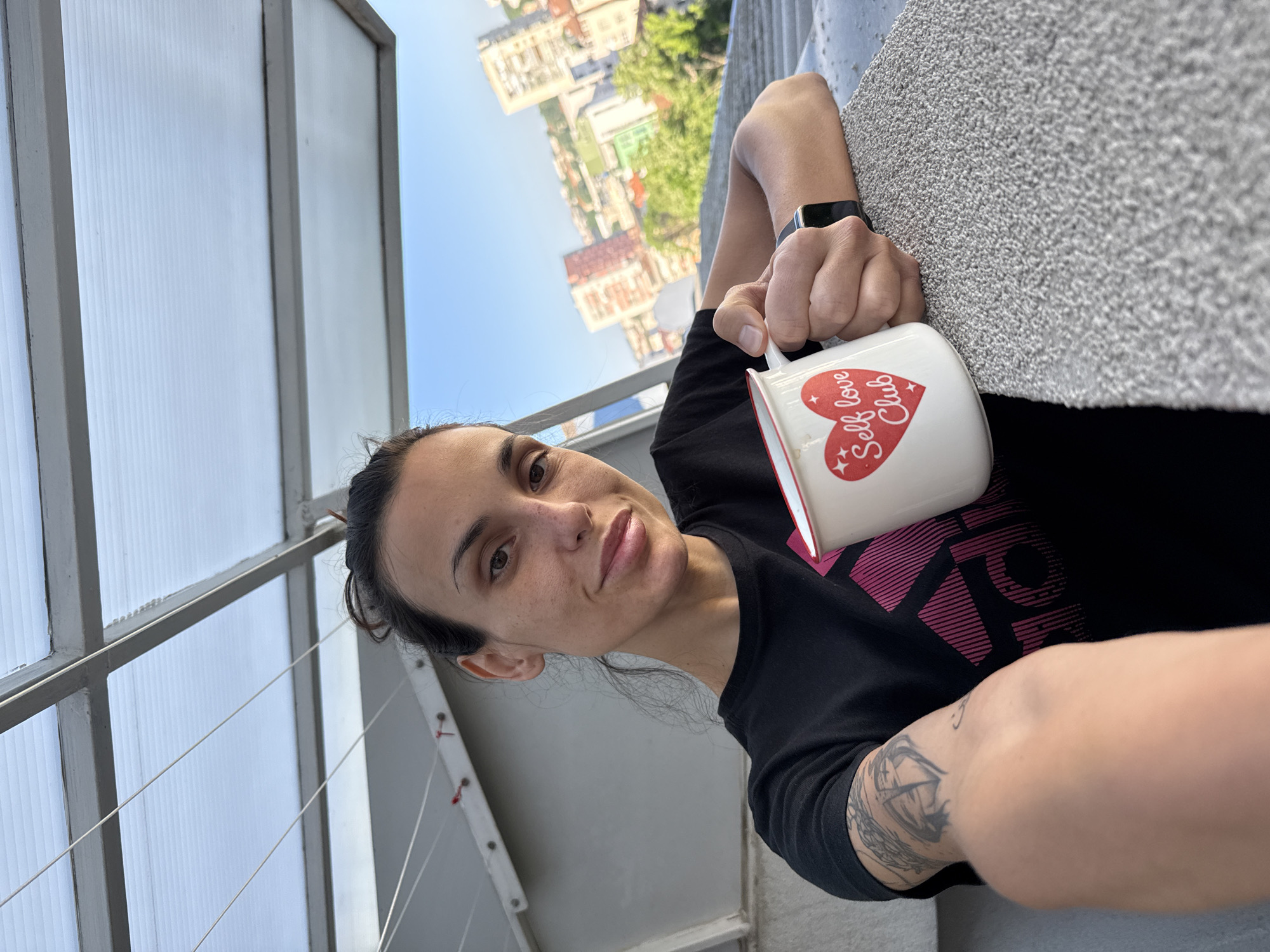 A woman with brown hair pulled back holds a white mug with a red heart and "Self Love Club" printed on it. She is wearing a black t-shirt with a magenta design. The woman has a tattoo on her left arm and is wearing a black smartwatch on her left wrist. She is on a balcony with a polycarbonate privacy screen. In the background is a cityscape with apartment buildings and trees. Based on architectural characteristics and the general layout of the city visible, the location is likely within the city of Pleven, Bulgaria.