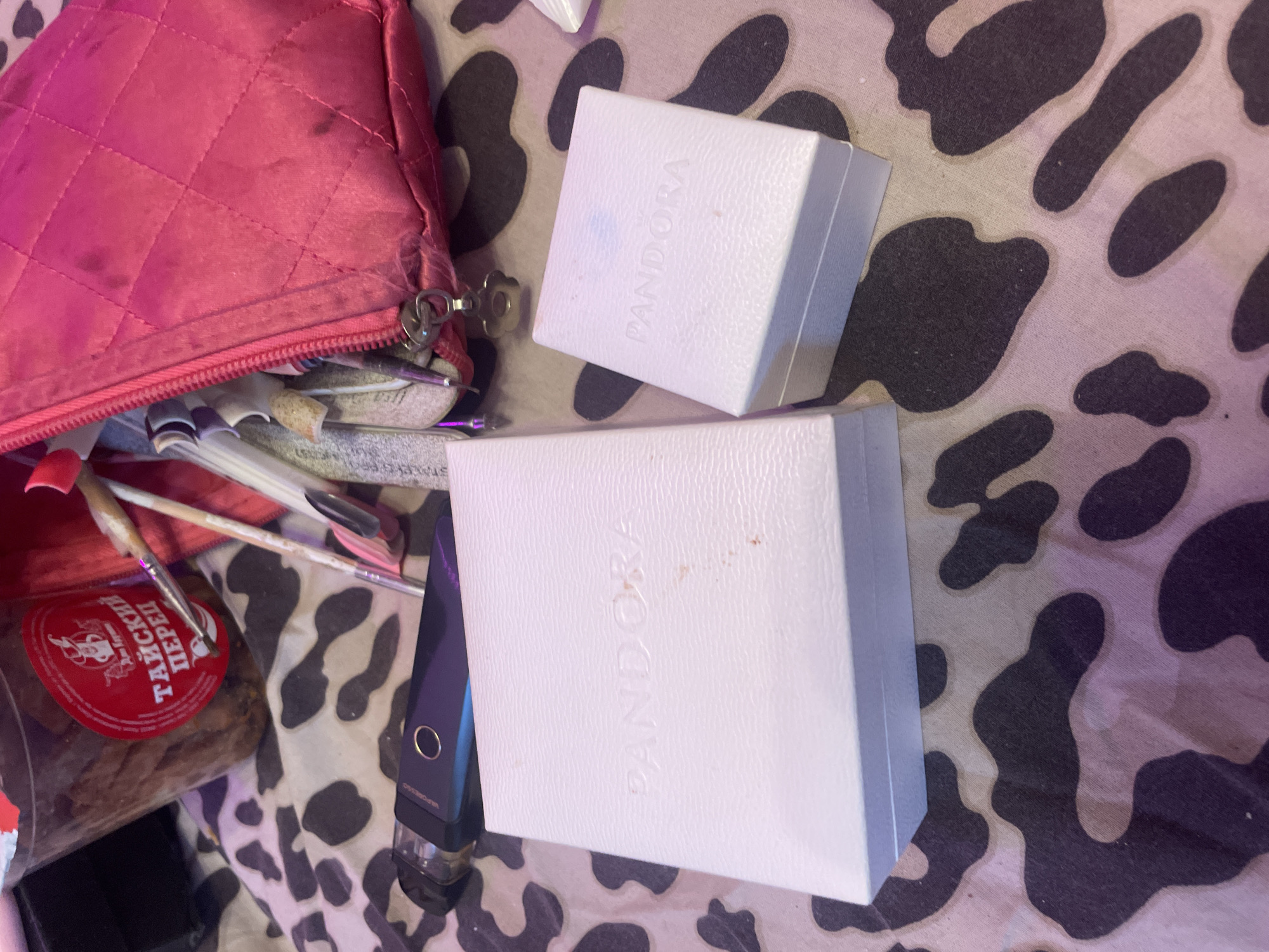 The image shows a close-up, overhead view of a bedside table in Ladozhskoe Ozero, Russia. On the table, there are two white Pandora jewelry boxes, one larger than the other, placed on a cow-print fabric. To the left of the boxes is a black vape pen, and behind it is a clear plastic container filled with food, featuring a red label with Russian text. To the right of the boxes, a bright pink, quilted cosmetic bag is partially open, revealing various items inside, including makeup brushes and nail files. The lighting is dim, suggesting it may be nighttime or indoors with artificial light.
