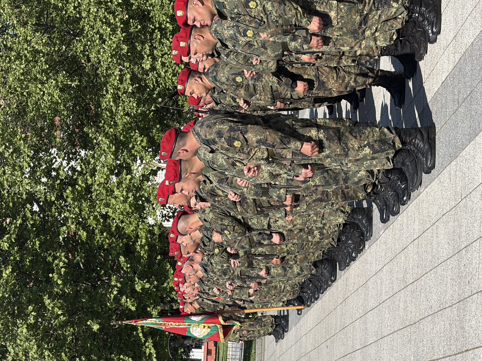 A group of soldiers in camouflage uniforms and red berets stand in formation on a paved area in Pleven, Bulgaria. They are lined up in rows, facing forward, with their hands clasped behind their backs. A flag with green, white, and red stripes is held by one of the soldiers at the front of the formation. A tree with green foliage is visible to the left of the soldiers. The paved area has horizontal lines. The scene appears to be a formal military event or ceremony.