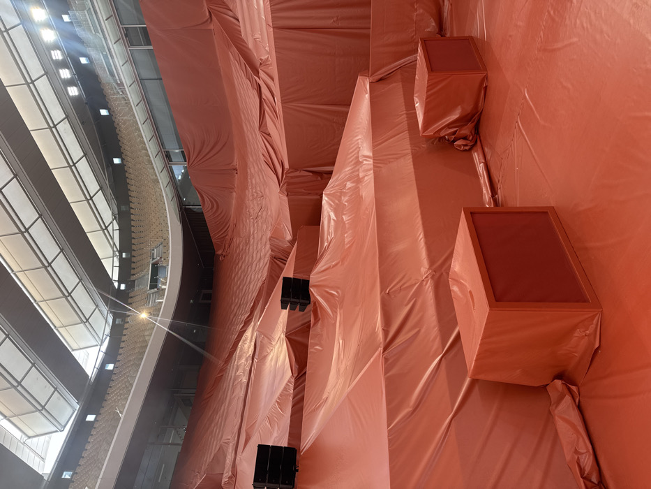 The scene depicts the interior of the Palais de Tokyo, located in Paris, France, largely enveloped by a vibrant orange, synthetic tarp material. This tarp meticulously drapes over multi-tiered architectural structures, floors, and various cubic objects, creating an undulating, wrinkled surface that dominates the visual field. Two distinct stacks of three black professional audio speakers are positioned on separate covered tiers, with a third, smaller, single speaker unit also covered by the orange tarp in the foreground. In the upper background, beyond the orange-covered sections, rows of light-colored, empty stadium-style seating are visible. The ceiling structure features translucent panels allowing ambient light, complemented by several visible artificial light fixtures, one of which produces a prominent lens flare. No individuals are present within the captured scene.