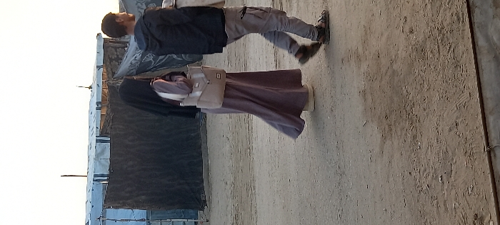 The scene, located in Khan Yunis, Palestinian Territory, depicts two individuals walking from left to right on an unpaved dirt path. On the left, a female figure wears a dark head covering (hijab), a long, light purple abaya-style garment, and light-colored platform sandals, carrying a beige shoulder bag on her right side. To her right and slightly ahead, a male figure walks, wearing a dark hooded jacket, light grey cargo pants, and dark sandals; he carries a light-colored, indeterminate object in his left hand. The ground is composed of loose, sandy soil with visible faint impressions. In the background, a series of makeshift shelters are constructed from various materials, including white, blue, and dark-colored tarpaulins draped over frames, consistent with a displacement camp environment. The sky above is pale and clear, indicating natural ambient light.