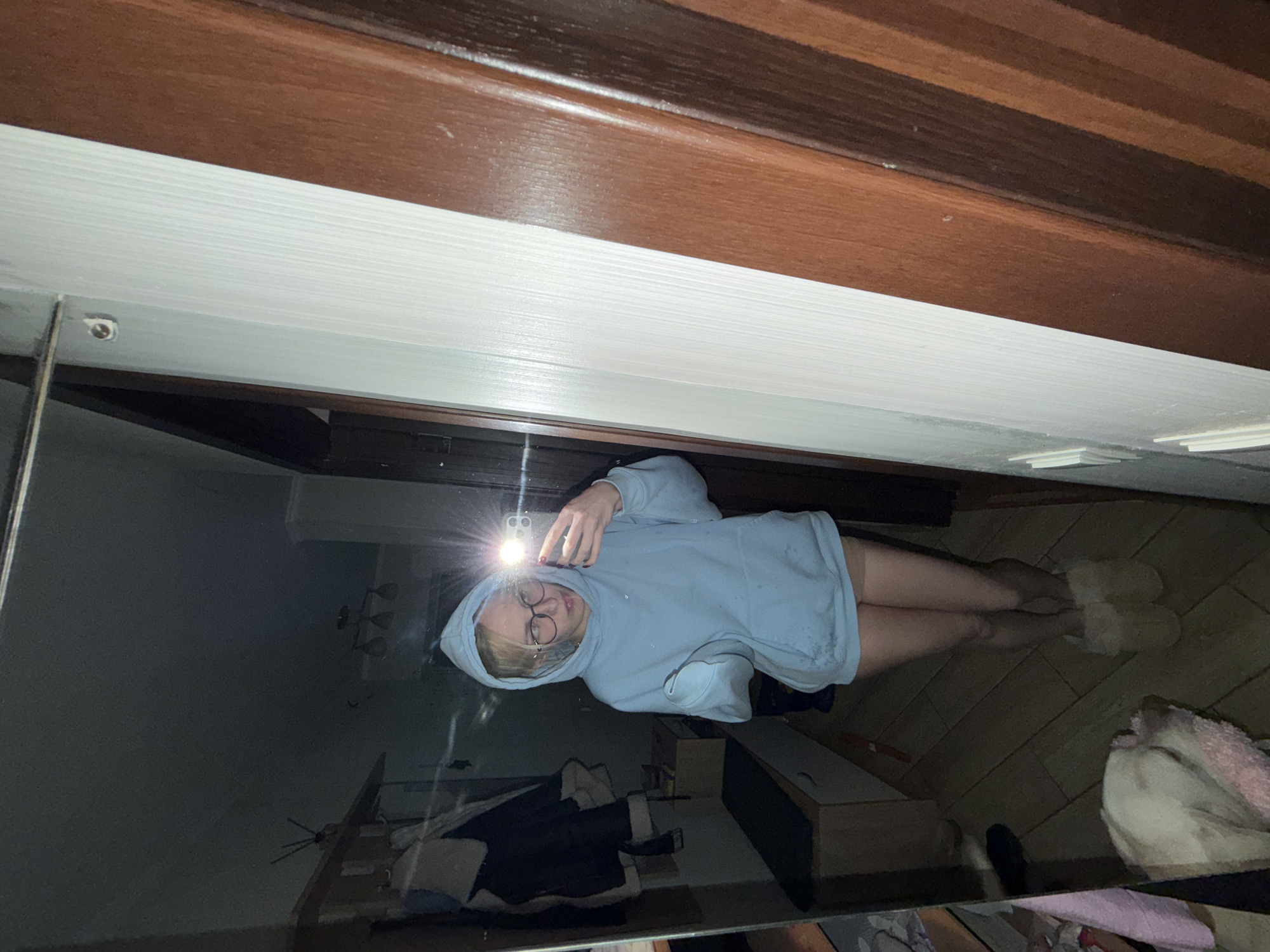 A young woman with blonde hair and glasses is taking a mirror selfie. She is wearing a light blue hoodie and light-colored slippers. She is standing in what appears to be a hallway or dressing area. In the reflection of the mirror, a dog is visible lying on a pink blanket. The environment appears to be a residential indoor setting. There are no discernible urban features that would place this image in Cheltuitori, Moldova.