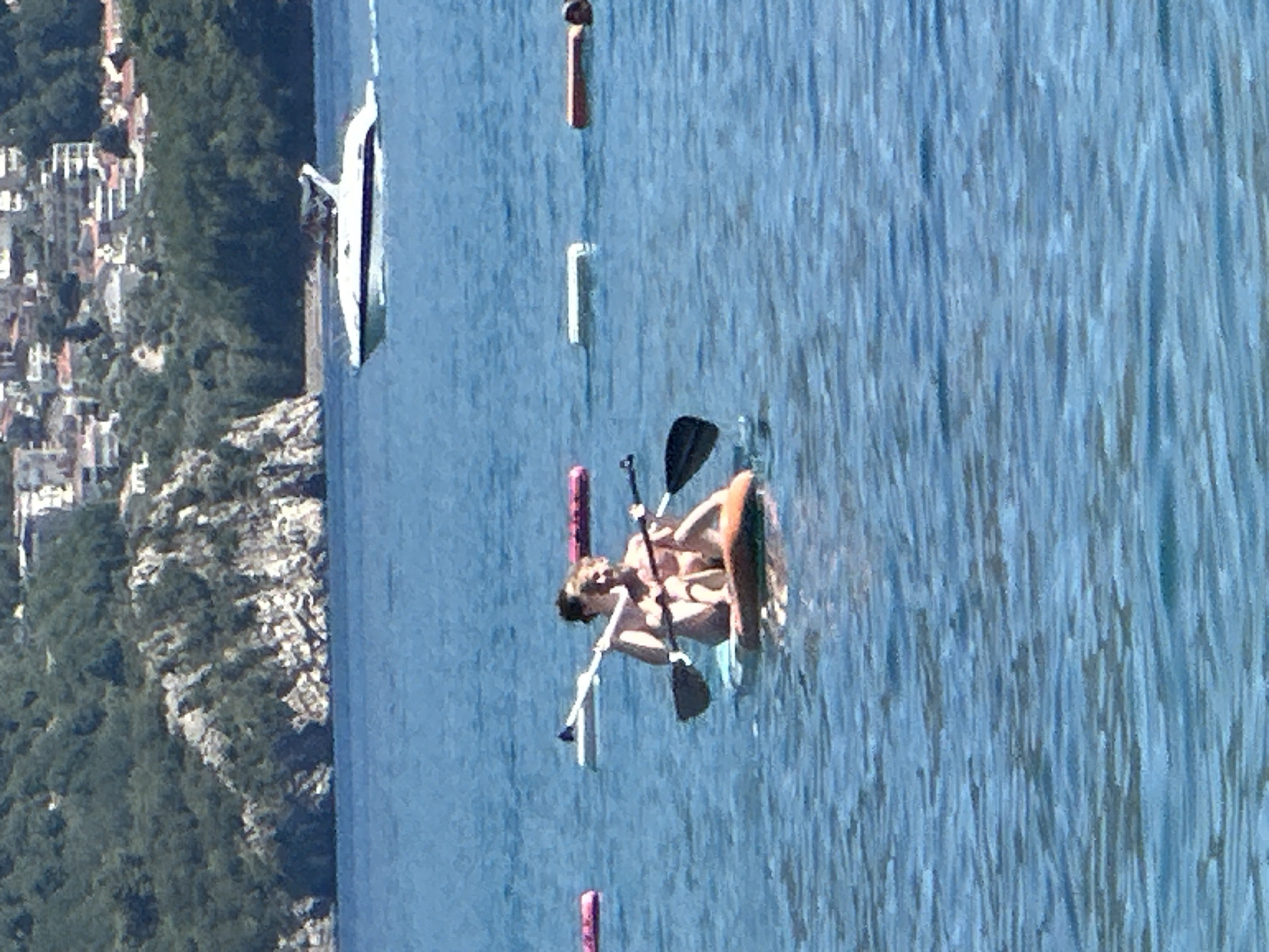 In Boreti, Montenegro, two individuals are seen on a paddleboard in the Adriatic Sea. The person in the front is holding a paddle, while the person behind them is also holding a paddle. They are both facing forward. The water is a clear blue, and there are a few buoys in the water. In the background, there is a coastline with lush green trees and buildings. A white speedboat is also visible in the distance. The scene appears to be a sunny day with calm water.