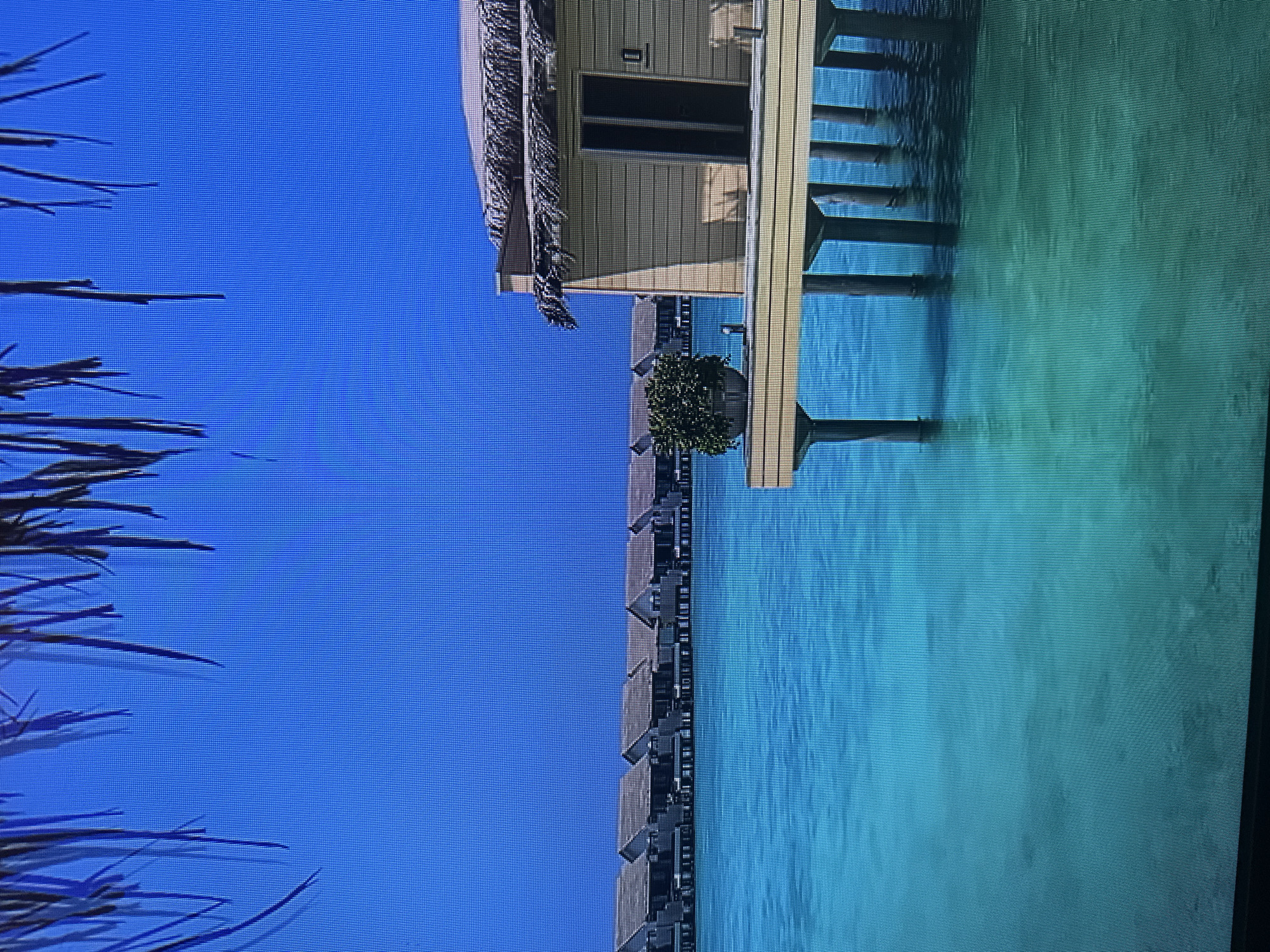 The image depicts a picturesque scene of overwater bungalows situated in a clear, turquoise body of water under a bright blue sky. In the foreground, palm fronds are visible, creating a natural framing effect. To the right, a single bungalow is prominently featured, elevated on stilts and featuring a thatched roof and light-colored siding. A potted plant sits on the deck of this bungalow. In the background, a row of identical bungalows stretches across the horizon. The water appears calm and shallow, with visible reflections of the bungalows and their supporting structures. There are no people or animals visible in the image. The overall impression is one of a serene, tropical resort. This scene is not located in Kyiv, Ukraine, as it depicts a tropical environment inconsistent with Ukraine's geography.