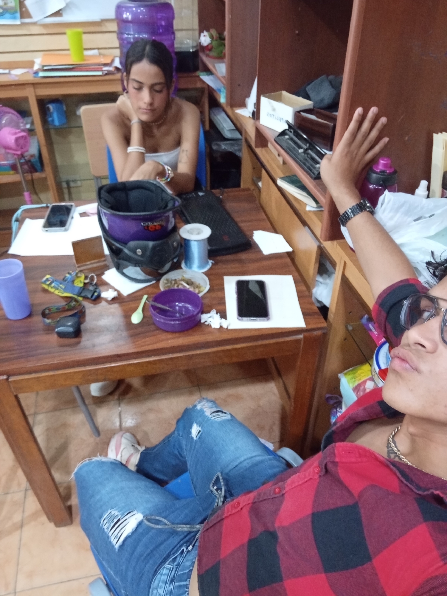 This image captures a casual indoor scene, likely in Juan Griego, Venezuela, featuring two young individuals. The male subject, in the foreground, appears to be taking a selfie. He wears a red and black plaid shirt, ripped blue jeans, and glasses, with a gold chain around his neck and a black watch on his left wrist. His arm is raised, holding the camera, as he looks towards it with a relaxed expression.
Seated across a wooden table from him is a young woman. She wears a white strapless top and multiple bracelets on both wrists. Her dark hair is pulled back, and she rests her chin on her hand, looking downwards with a pensive or slightly bored expression.
The wooden table between them is quite cluttered. Prominently displayed are a black and purple motorcycle helmet, which has partially visible text resembling "Giro" or "Gira," and two smartphones (one black, one silver) resting on white papers. Other items include a blue spool of thread or wire, a small bowl with food remnants, a purple bowl with a green spoon, a purple plastic cup, a small brown rectangular object, yellow and blue sandals, and a black strap device. A pink oscillating fan and a laptop are also visible on the table further back.
Behind the female subject and extending along the wall are wooden shelves, filled with various miscellaneous items, including boxes, a large purple water jug, and a purple water bottle. The floor is light brown tiled. The overall impression is a relaxed, everyday setting, possibly a home or communal space, during daytime hours.