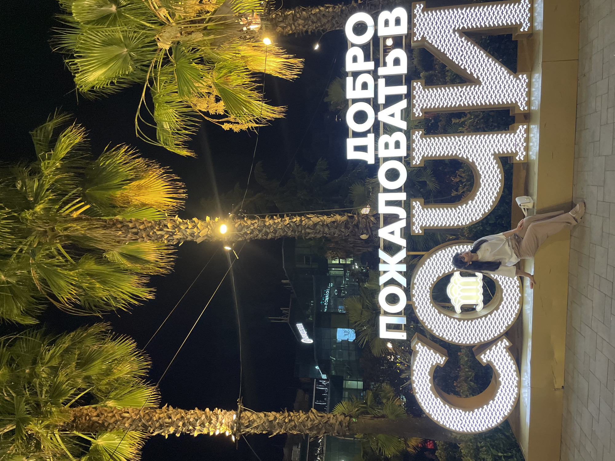 A woman is posing in front of a large, illuminated sign that reads "ДОБРО ПОЖАЛОВАТЬ В СОЧИ" which translates to "Welcome to Sochi." The sign is made of large, white, block letters that are outlined with a decorative pattern of small, white shapes. The letters of "СОЧИ" are particularly large and are mounted on a golden platform. The woman, wearing a white long-sleeved shirt and light brown pants, is sitting on the edge of this platform, looking to her right. She is wearing a white purse and tan shoes. The sign and the woman are illuminated by the bright lights of the sign itself, as well as by streetlights and lights from nearby buildings. The scene is set at night, with palm trees visible in the foreground, suggesting a warm climate. In the background, there are several buildings with glowing windows and signs, including one that appears to be a hotel with the word "ОРИОН" visible. The location is likely a public square or promenade in the city of Sochi, Russia, known for its resort destinations and palm trees.