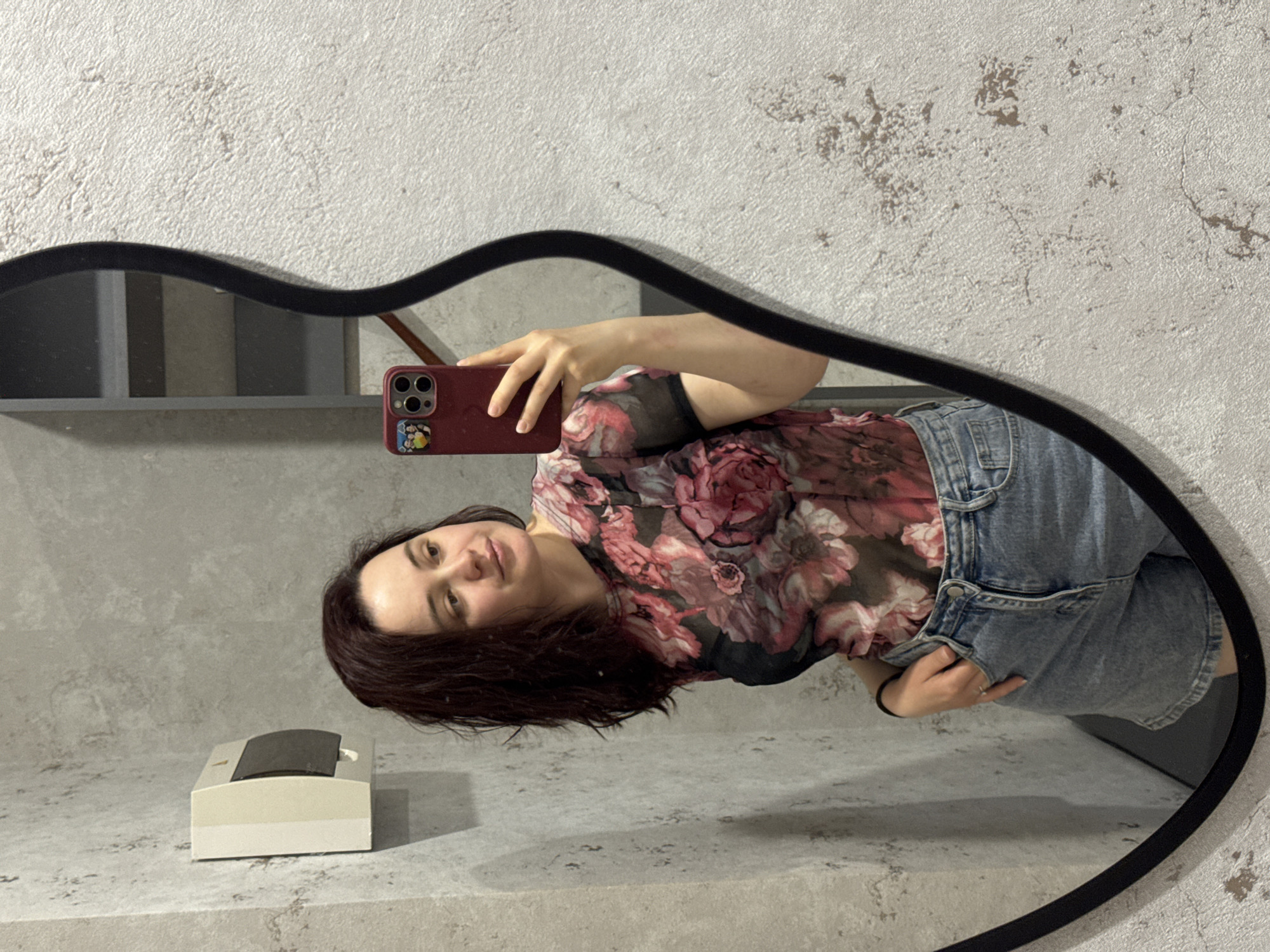 A woman stands in front of a black-framed, amoeba-shaped mirror, holding a red smartphone in her right hand to take a selfie. She is dressed in a semi-sheer floral print top, exposing a black camisole underneath, and a light blue denim skirt. The location appears to be indoors, with a gray and white textured wall in the background. To the left of the mirror's reflection is a beige object. The mirror reflects a gray shelf with some items on it. The selfie depicts the woman looking at the camera with a slight smile. There is no specific information confirming this is Izhevsk, Russia.