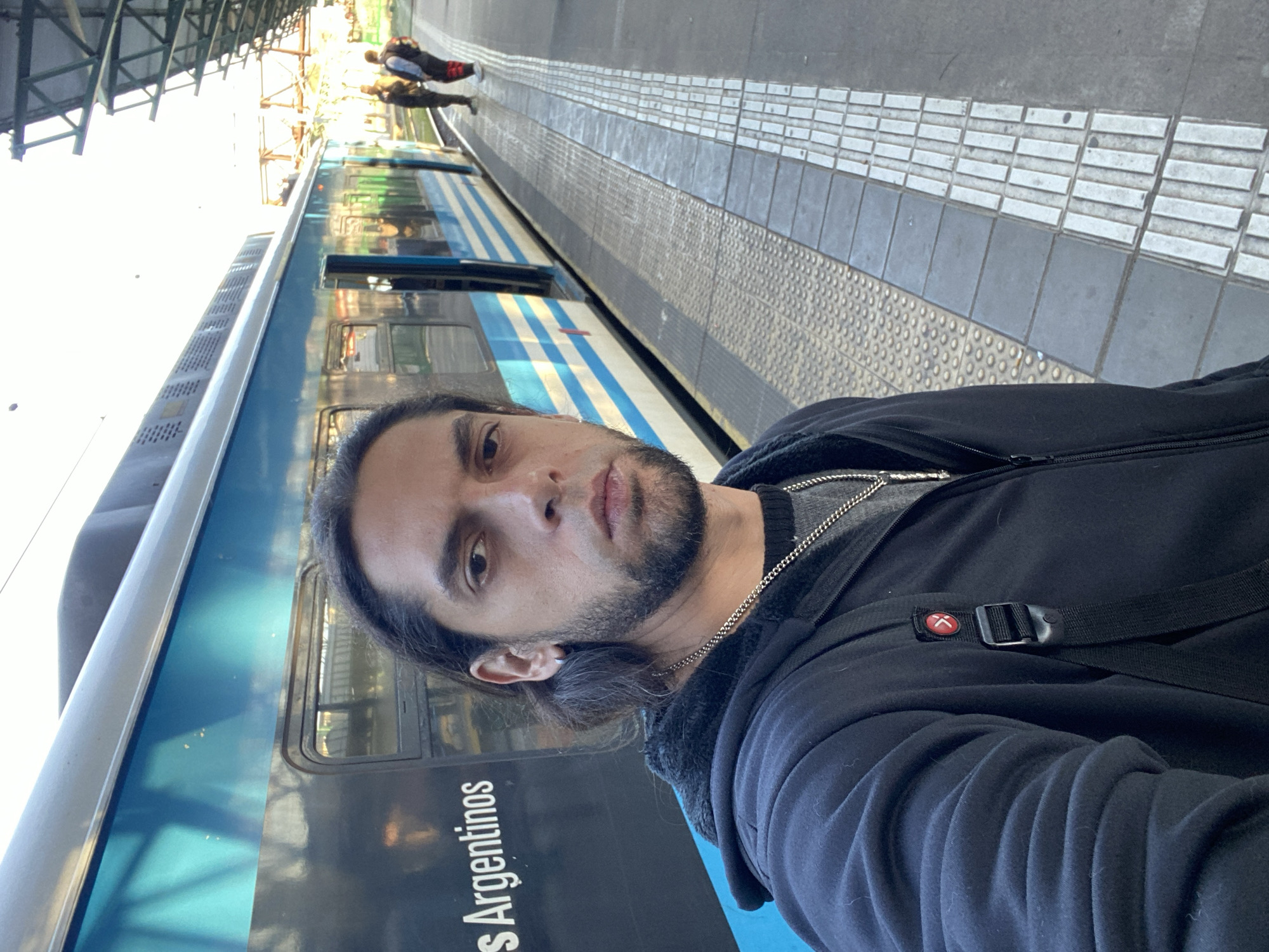 A man with long dark hair and a beard stands on a train platform. He is wearing a black jacket and a silver chain. Behind him is a train with the words "s Argentinos" visible on the side. The platform has a textured edge, and there are other people in the background, further down the platform. The image appears to be taken in a train station in Buenos Aires, Argentina.