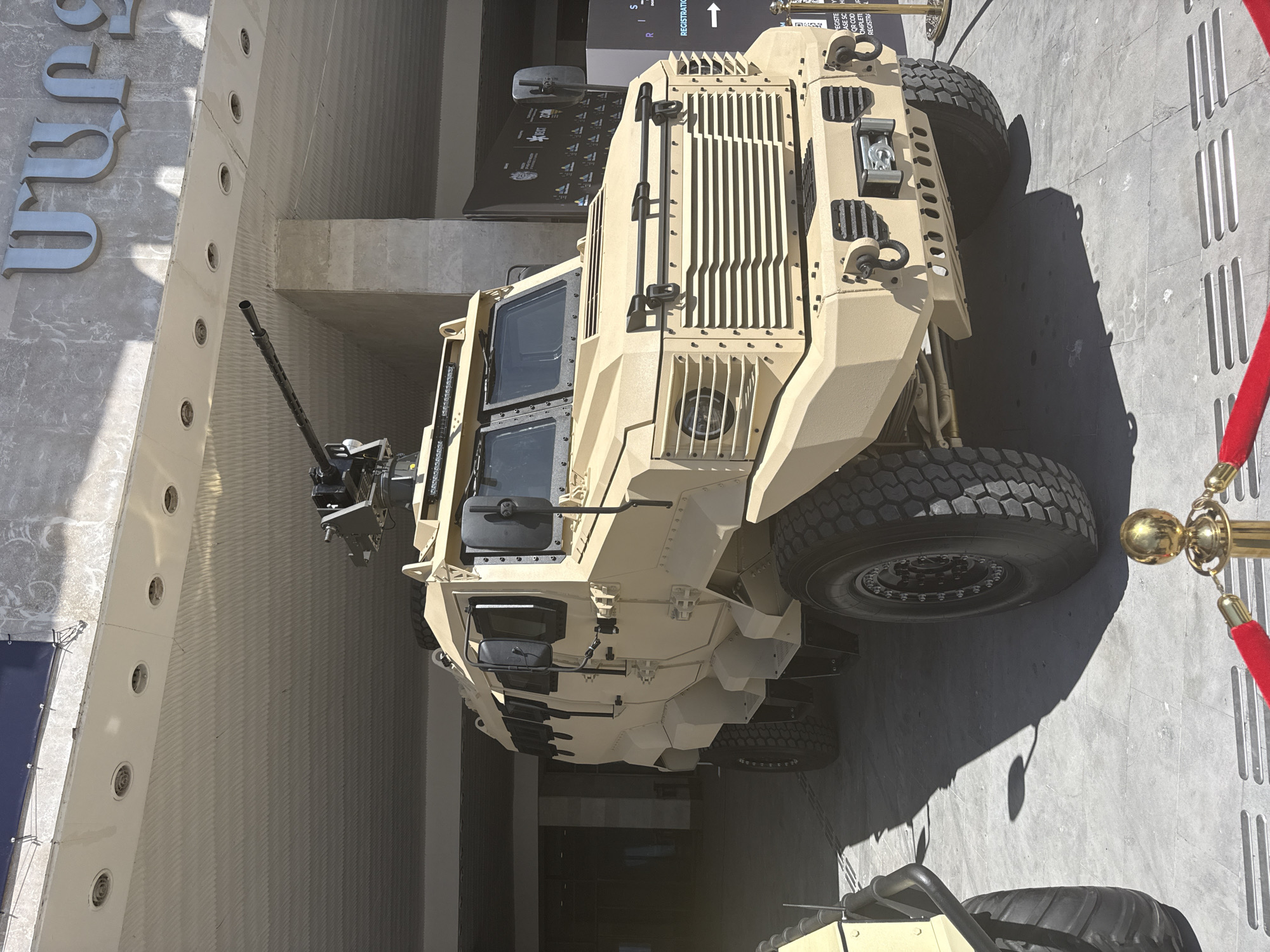 A tan-colored, heavily armored military vehicle is the central subject, prominently displayed outdoors under bright, sunny conditions. The vehicle appears to be an infantry mobility vehicle or an armored personnel carrier, characterized by its angular design, robust construction, and large, rugged off-road tires. A weapon, likely a heavy machine gun, is mounted on a turret on its roof, facing towards the left side of the frame. The vehicle's windows are armored, and its exterior features several protective grilles and fixtures.
It is parked on a paved, light gray ground, which shows sharp, dark shadows cast by the vehicle and other elements, indicating a sunny day, likely mid-day or early afternoon. To the right, a red velvet rope barrier supported by ornate gold stanchions suggests an organized display or event area.
In the background, a modern building facade is visible, featuring a textured, light gray surface. Large, metallic, ornate letters are affixed to the building, with visible characters suggesting Armenian script, specifically "ՌԱՆ". Further back, a dark sign with white text is partially visible, clearly showing "REGISTRATION" and an upward-pointing arrow, along with fragments of other words like "CASE SC" and "COMPLET". No people are visible in the scene. The overall context suggests the vehicle is part of an exhibition, defense expo, or similar public display event.