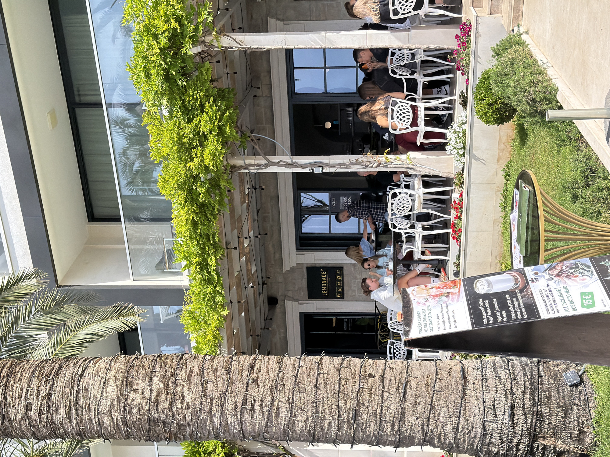 The image captures an outdoor cafe scene, likely in Bečići, Montenegro. A palm tree trunk dominates the foreground, partially obscuring the view. The cafe, named "Lemonade," features white ornate chairs occupied by patrons. Some are seated and interacting, while others are possibly awaiting service. The cafe's facade includes large windows and hanging greenery. A menu board displays breakfast and drink options in multiple languages, suggesting a tourist-oriented establishment. The environment includes greenery, indicating a pleasant outdoor setting.