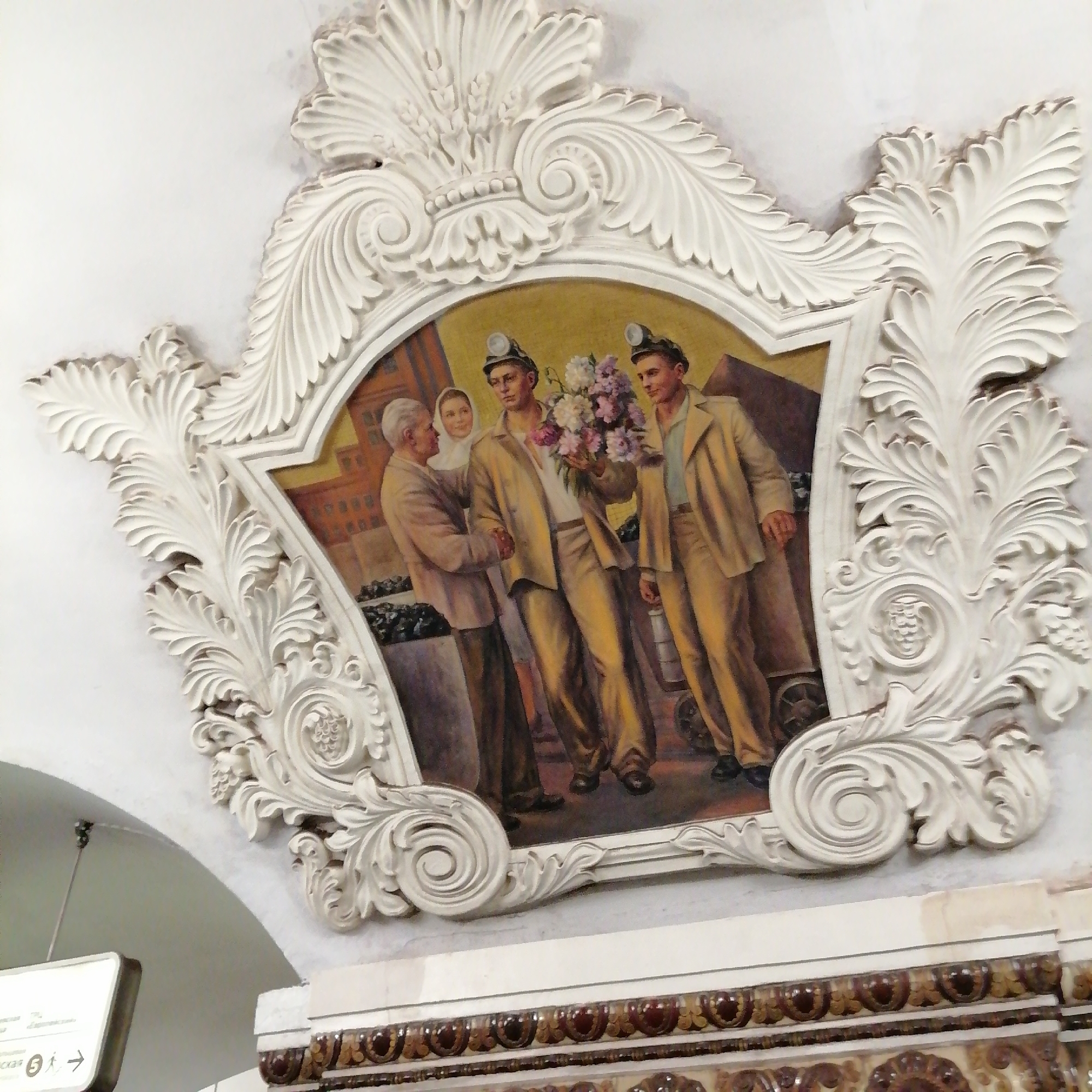 A mosaic depicting people with coal mining helmets on, surrounded by an ornate white decorative frame, sits above a brown and gold patterned wall. The mosaic features two men in miner's helmets holding a bouquet of flowers, an older man extending his hand in greeting, and a woman standing behind him. The mosaic is likely located in a Moscow metro station as many stations are decorated with socialist-realist art.