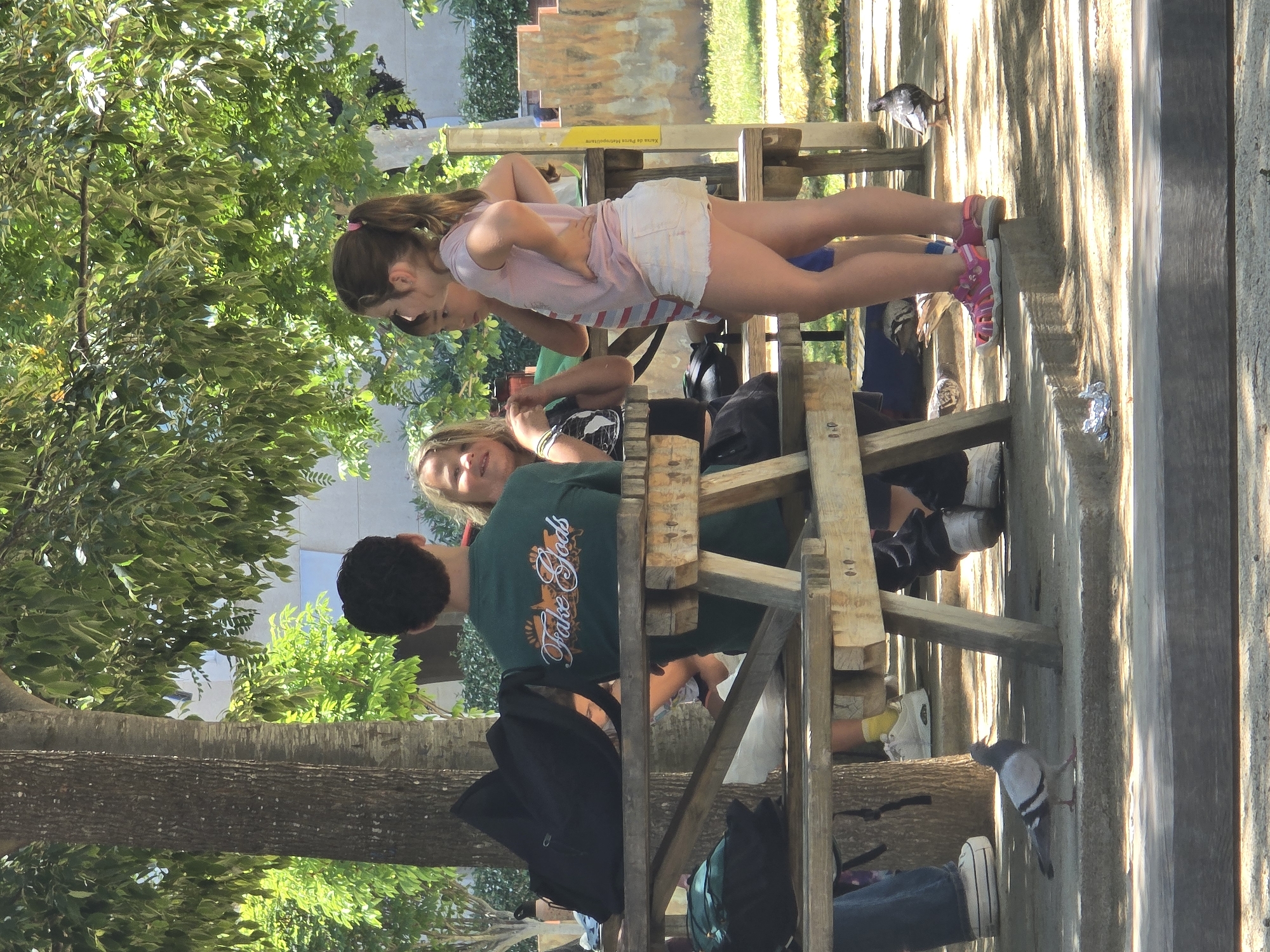 A group of people, including at least three young girls and one young man, are gathered around a wooden picnic table outdoors in Cornellà de Llobregat, Spain. The young man, wearing a green t-shirt, is seated. One of the girls, wearing a pink t-shirt and white shorts, is standing and facing away from the camera. Another girl, with blonde hair, is also seated and appears to be interacting with the young man. A black backpack rests on the table, and another backpack is on the ground. The environment appears to be a park or recreational area, with trees and sunlight filtering through the leaves. Two pigeons are visible on the ground near the table.