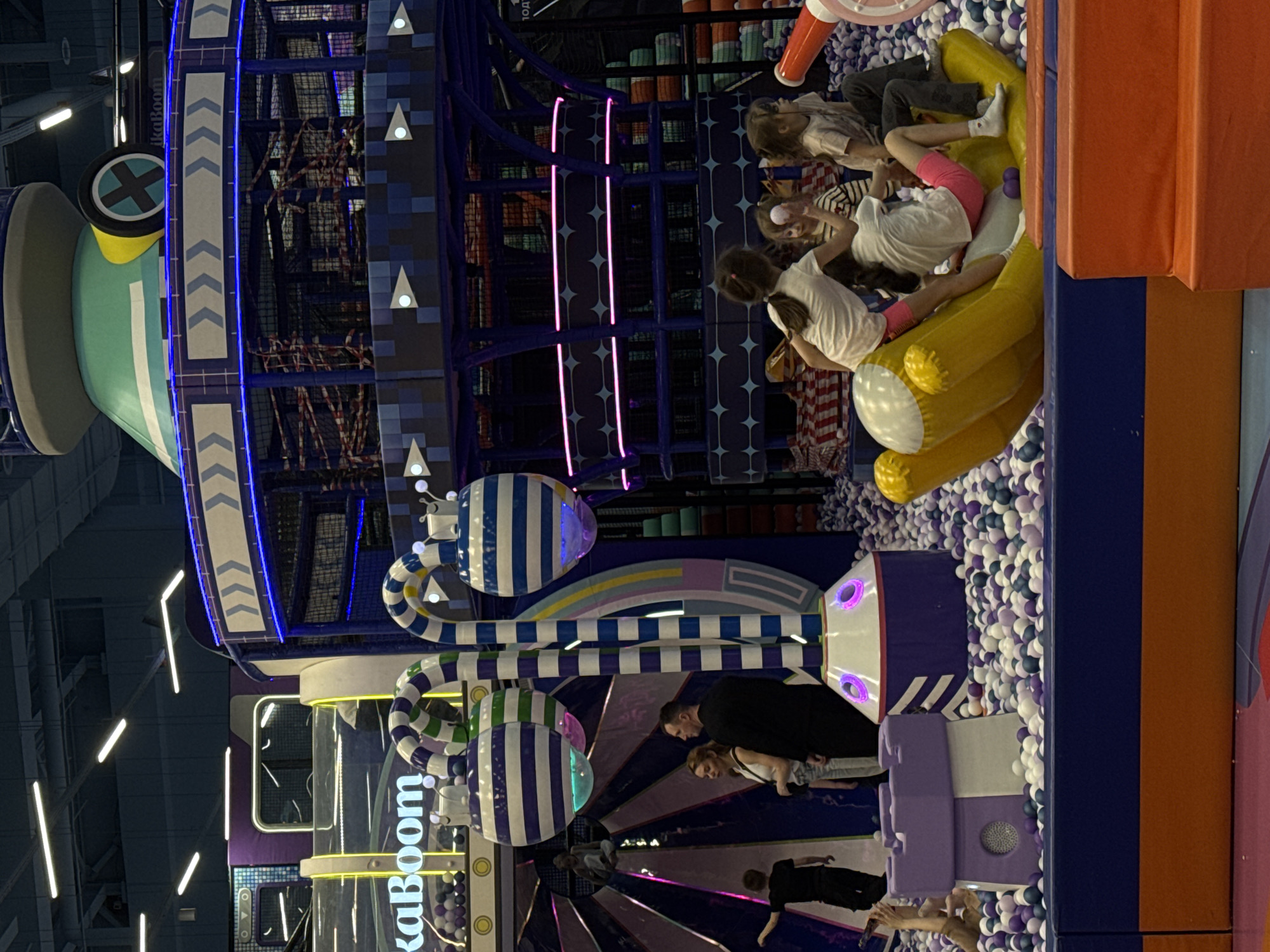 This image depicts a lively indoor children's play area, likely an entertainment center or amusement park in Saint Petersburg, Russia. The scene is filled with vibrant colors and various play structures.
Several children are actively engaged in play. A group of children are in a large ball pit filled with white, purple, and light blue plastic balls, with some climbing on a large, yellow inflatable structure that resembles a boat or raft. Other children are visible within and around the multi-level play structures. A few adults are also present, seemingly supervising or accompanying the children. One adult male and one adult female are visible standing near a striped, cylindrical structure.
The dominant feature is an elaborate, multi-level play structure with blue and purple scaffolding, enclosed by netting. Sections of this structure are illuminated by bright blue and pink/purple neon lights, creating a dynamic visual effect. Prominent decorative elements include large blue and white striped cylinders and arches, as well as a large green, white, and yellow funnel-like structure topped with a black "X" on a yellow circular emblem. The words "KaBoom" are clearly visible in white lettering on several parts of the play equipment, identifying the attraction or venue. The overall atmosphere is energetic and playful, characteristic of a children's activity center. The lighting suggests an indoor setting, with artificial illumination creating a bright environment for play.