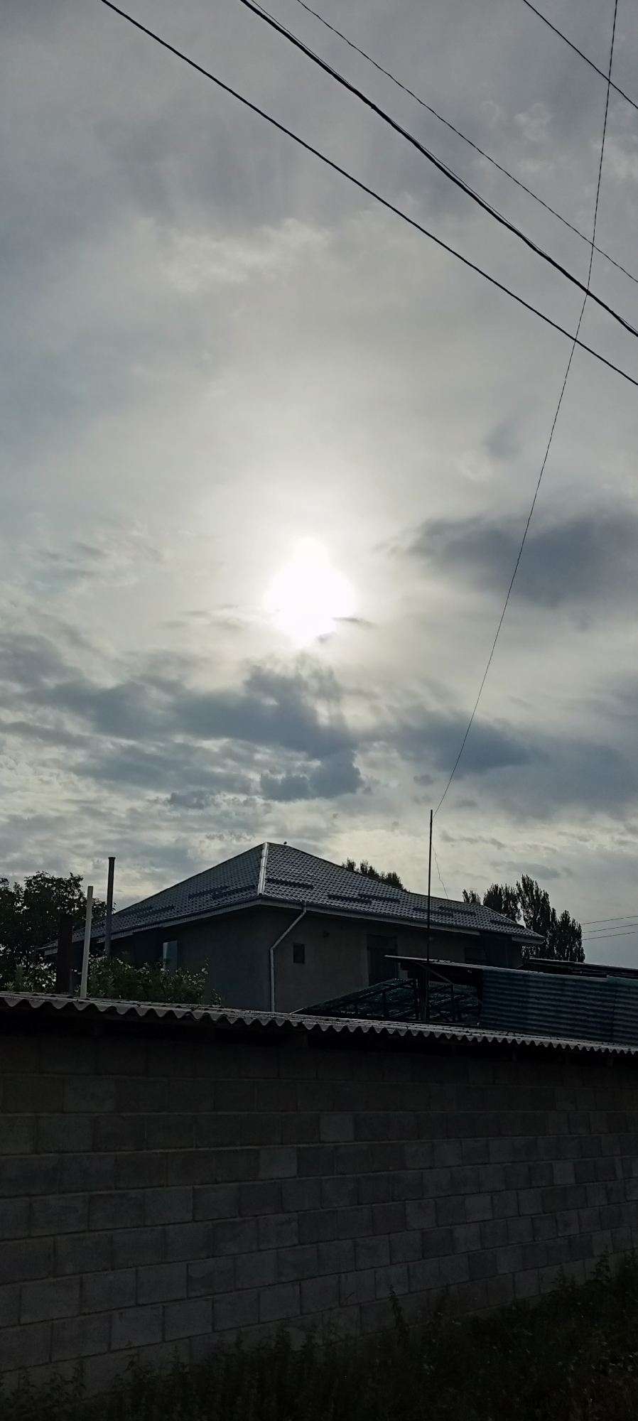In Sokuluk, Kyrgyzstan, a cloudy sky dominates the upper portion of the image, with the sun partially obscured by clouds, casting a bright glow. Several dark, thin wires stretch across the sky, crisscrossing and running at various angles. Below the sky, a building with a dark, sloped roof is visible. The building appears to be under construction or recently built, with a plain, grey exterior wall. A small chimney or pipe protrudes from the roof on the left side. In front of the building, a corrugated metal structure and a brick wall are present, partially obscuring the lower part of the building and the surrounding vegetation. The brick wall, constructed from rectangular blocks, runs horizontally across the bottom of the image. Dark green foliage and grass are visible at the base of the brick wall and around the building. The scene appears to be an outdoor residential or semi-rural area.