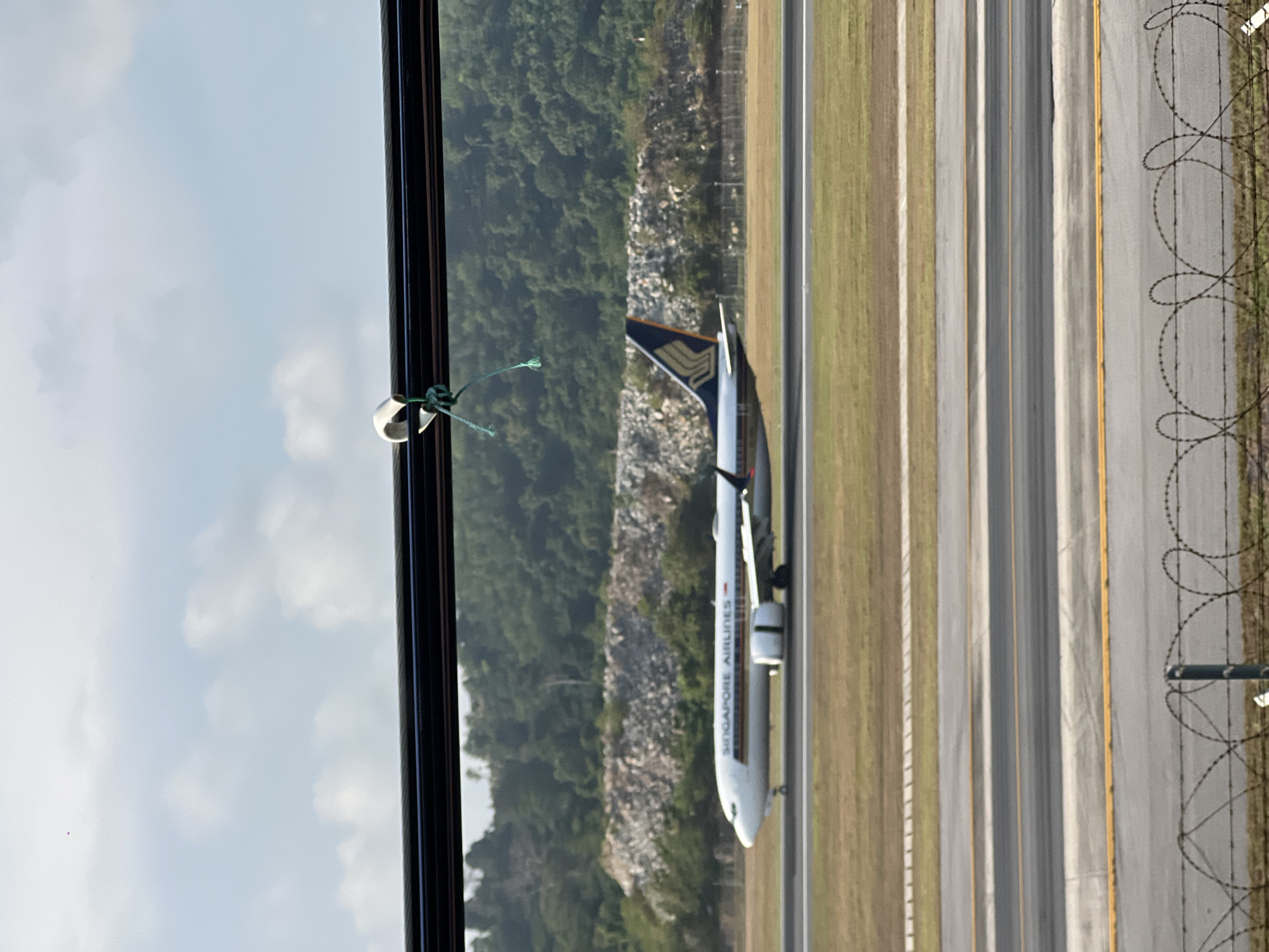 A sideways-oriented view from a window, showcasing a Singapore Airlines airplane positioned on an airport runway. The runway stretches along the right side of the image, with a section of grassy land separating it from a barbed-wire fence. Beyond the fence, another strip of land is visible. The left side of the image reveals a view of lush greenery, and behind the trees, some rocky terrain can be seen. The sky, filled with scattered clouds, is visible in the top-left corner. The location is possibly near Tambon Mai Khao, Thailand, given the surroundings.