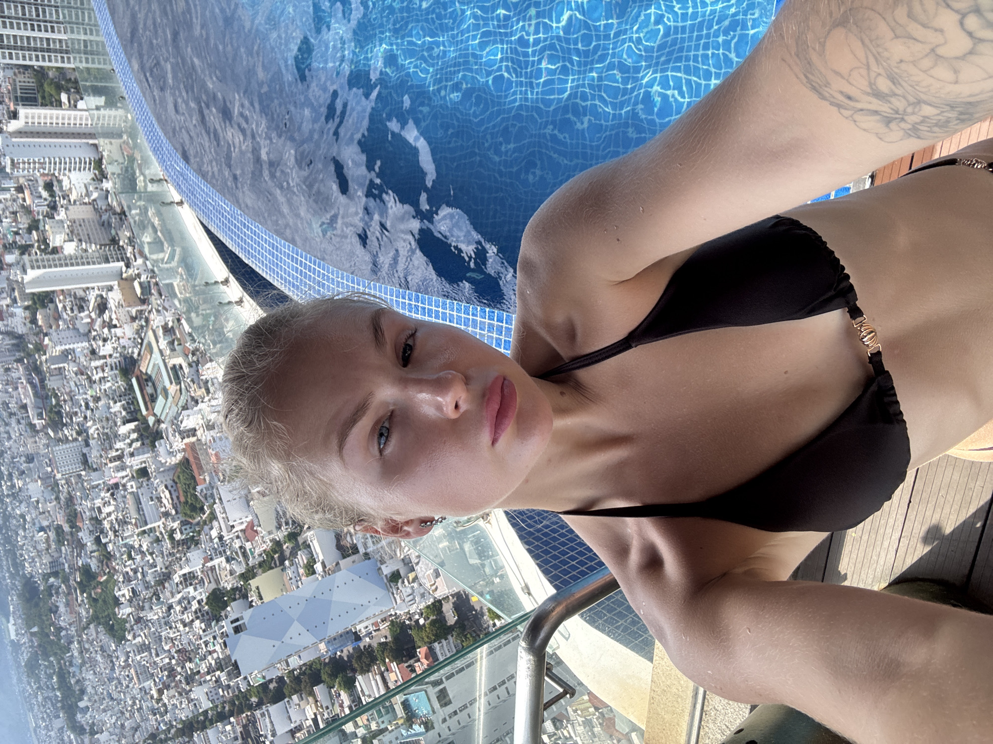 This media file is a portrait of a person in a black bikini, taken from a high vantage point, likely a rooftop pool. The person, with blonde hair and light skin, is looking directly at the camera with a neutral expression. They have a tattoo on their right arm.
The setting is a bright, sunny outdoor environment, suggesting daytime. The person is positioned next to a swimming pool with clear blue water and a tiled edge. In the background, a sprawling cityscape is visible, characterized by numerous buildings of varying heights and architectural styles, with lush green trees interspersed throughout. The sheer scale of the city below indicates a densely populated urban area. A railing, likely part of the pool area or observation deck, is visible in the foreground.
The image appears to be a selfie, given the angle and the inclusion of the photographer's body. The overall mood is relaxed and possibly celebratory, given the swimwear and the luxurious setting. There are no visible text elements in the image. The context suggests this is likely a vacation or leisure activity in a warm climate.