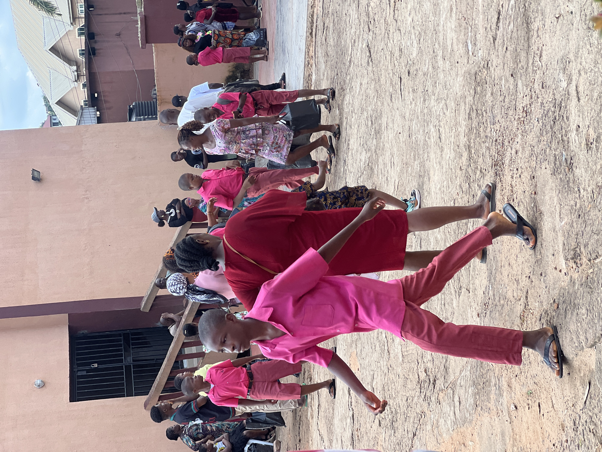 In Nnewi, Nigeria, a group of people, primarily children and young adults, are gathered outdoors. The scene appears to be in front of a building with a peach-colored exterior and dark-barred windows. A wooden railing and steps are visible to the left of the frame. The ground is a mixture of dirt and small stones.
Many of the individuals are wearing vibrant pink or red clothing. A young boy in the foreground is wearing a bright pink short-sleeved collared shirt and red knee-length shorts. He is wearing black flip-flops and appears to be walking away from the viewer. To his right, a young woman in a floral dress is carrying a black plastic bag. Behind them, other individuals in similar colored attire are visible, along with adults in various casual clothing. Some individuals are holding bags. The sky above is partly cloudy. The overall impression is of a gathering or event taking place in a public outdoor space.