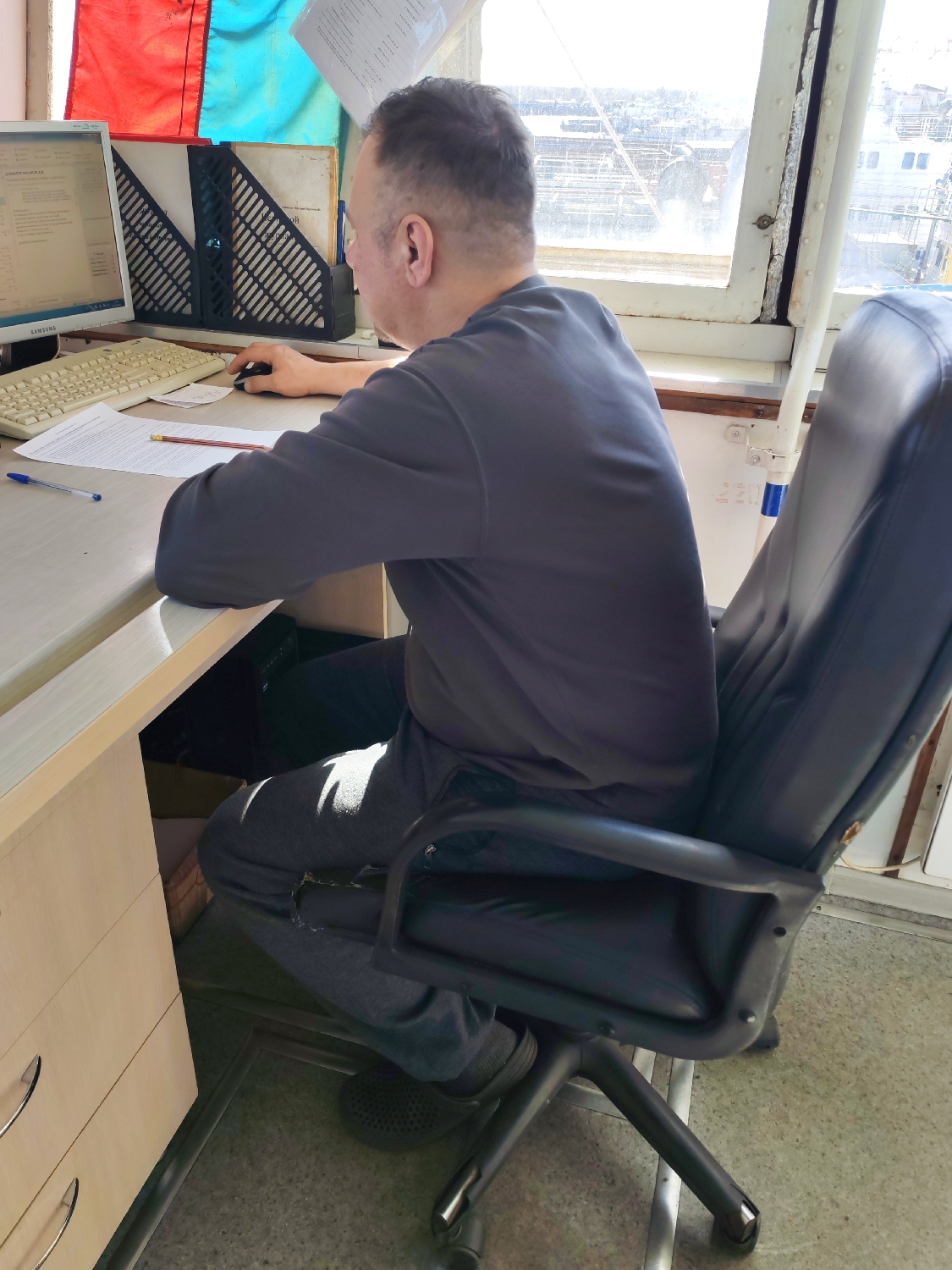 A man is seated in a black, rolling office chair at a light-colored wooden desk, with his back mostly to the viewer. He has short, dark hair and is wearing a dark grey long-sleeved shirt and grey sweatpants. His left hand rests on a computer mouse, while his right arm is on the desk. He appears to be focused on the computer screen in front of him.
On the desk are a beige computer keyboard, a white Samsung monitor, a mouse, several white papers with text, a blue pen, and a pencil. To the left of the monitor, a black document holder with a grid pattern contains papers, and a small black electronic device is also present.
Behind the desk and the man is a large, multi-paned window, letting in bright daylight that suggests it is a sunny day. Through the window, a maritime or industrial scene is visible, likely a ship or structures in a port area. Partially visible flags, one red and one light blue, hang above the window. The window frame appears sturdy and somewhat weathered.
Visible text includes "SAMSUNG" on the computer monitor. Partially visible Cyrillic text on the screen reads "ДОКУМЕНТЫ ДЛЯ ЗАГРУЗКИ" (Documents for loading/downloading). Other Cyrillic text is visible on documents held in the black holder. The man's grey sweatpants have a visible tear on the right knee. He is wearing dark, slip-on shoes.
The setting appears to be a functional workspace, possibly within a ship's bridge or a port office, given the view and flags. The man is engaged in administrative or data entry work on the computer, surrounded by documents.