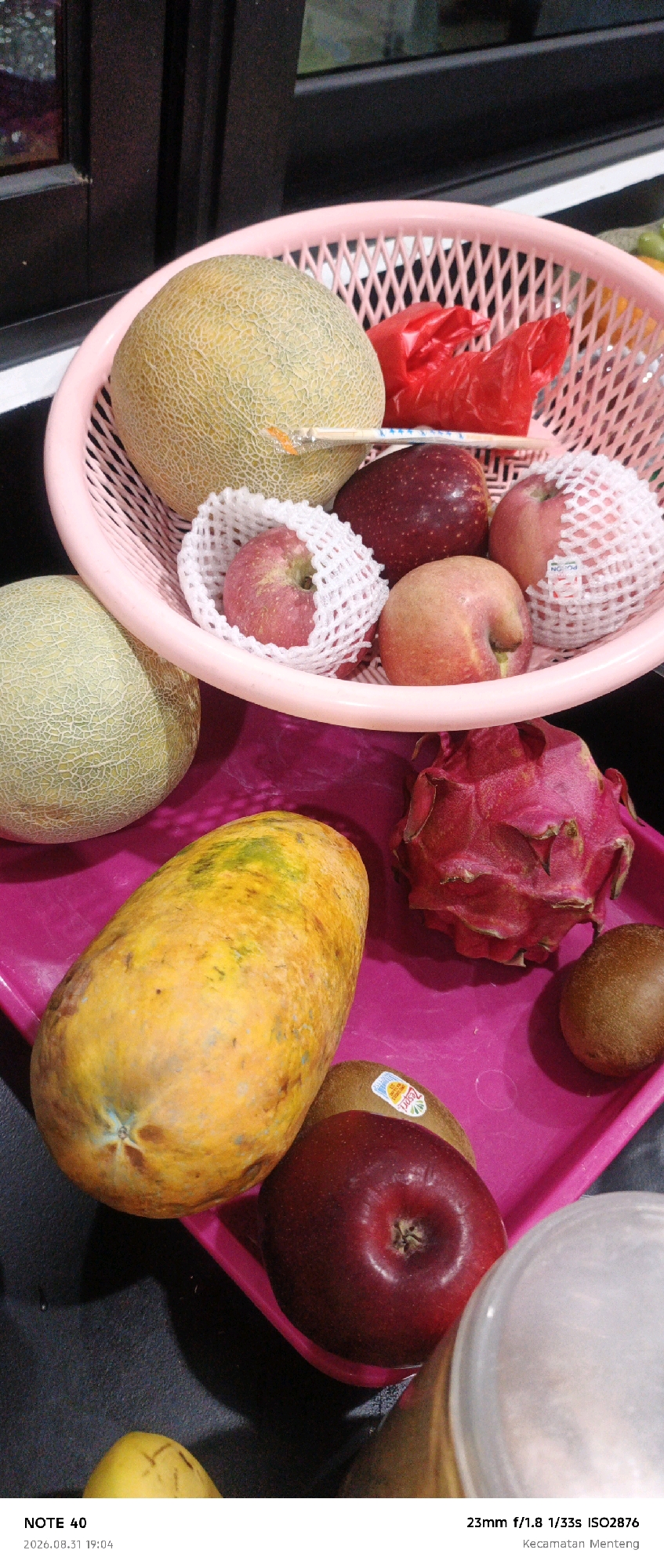 A still life photograph displays a variety of fruits arranged in a pink plastic basket and on a pink tray. In the basket, a large cantaloupe sits next to a red plastic bag. Several apples are also present, some individually wrapped in white foam netting. On the pink tray, a ripe papaya is positioned alongside a dragon fruit, two kiwis, and a dark red apple. The scene is lit, creating shadows. A text overlay at the bottom indicates "NOTE 40" and a date and time "2026.08.31 19:04", along with camera settings "23mm f/1.8 1/33s ISO2876" and the location "Kecamatan Menteng". Kecamatan Menteng is a district in Central Jakarta, Indonesia.