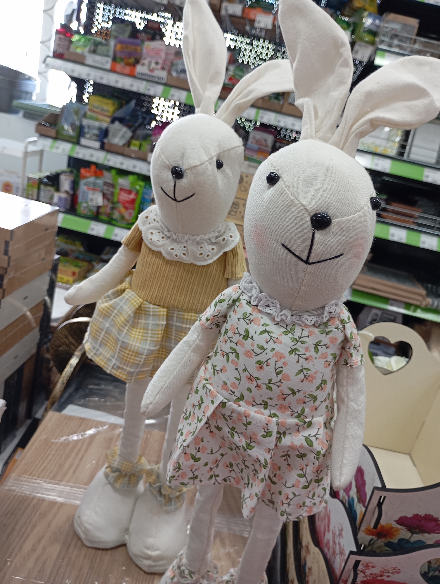 Two stuffed bunny dolls stand on a wooden surface inside a retail store. The bunny on the left wears a yellow dress with a white lace collar and a yellow and grey plaid skirt, while the bunny on the right wears a white dress with pink floral patterns and a white lace collar. Both bunnies have white ears and black button eyes and noses. The store shelves are visible in the background, stocked with various products. The location is Unknown, Unknown.