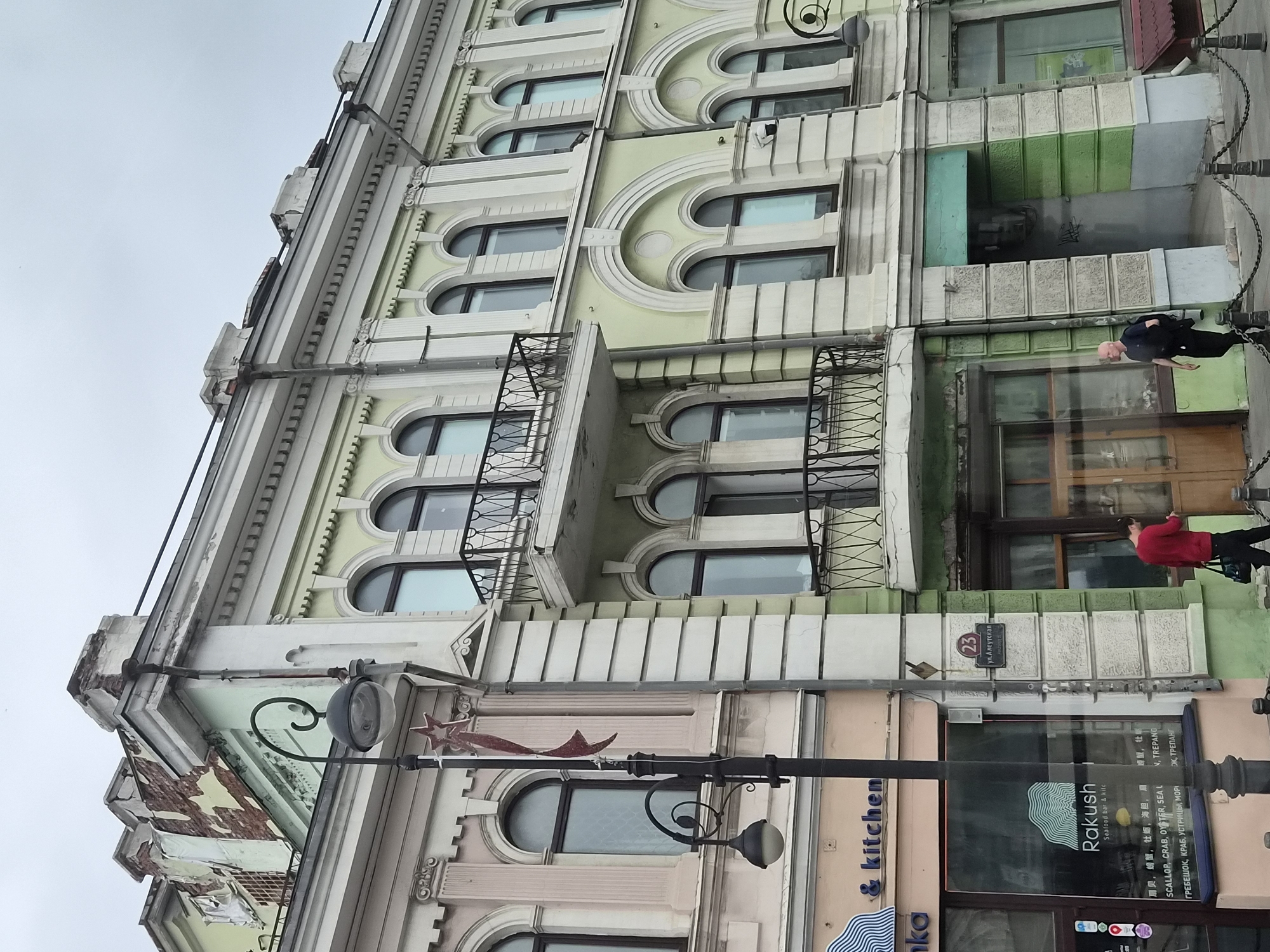 A medium-shot image shows the exterior of a multi-story building in Vladivostok, Russia. The building's facade is painted in a light green and beige color scheme, with architectural details like columns and arched windows. The upper part of the building shows signs of damage and disrepair, with exposed brickwork and peeling paint. A black lamppost with a decorative curved arm stands in the foreground, adorned with a red star ornament. The ground level features storefronts, including one with a sign that reads "Rakush Seafood Bar & Kitc". Two men are visible on the sidewalk in front of the building; one in a red sweater is walking away from the camera, and another in a dark shirt and pants is walking towards the camera. The sky is overcast and grey. The street is paved and lined with a metal chain fence.