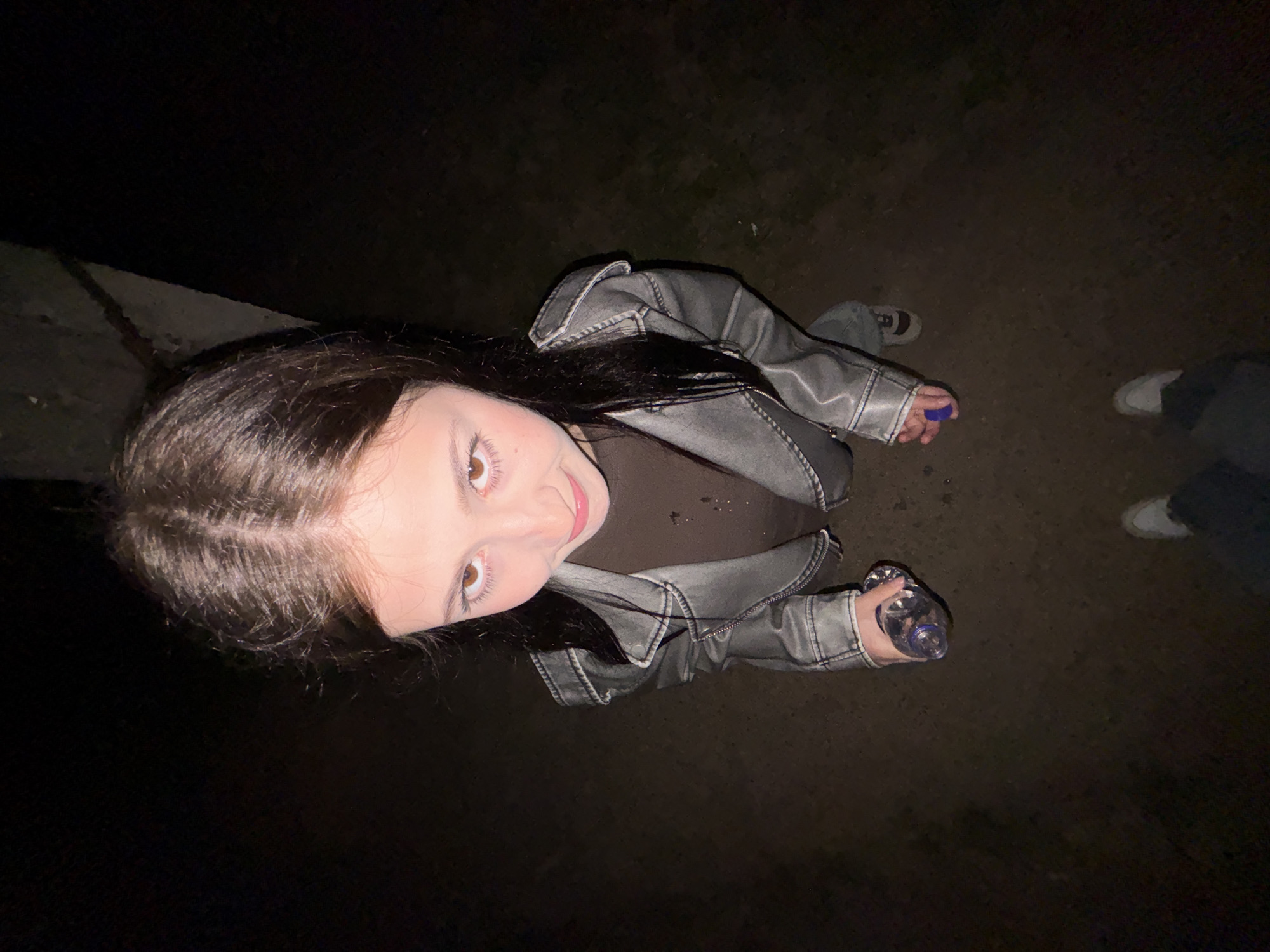 A high-angle shot shows a young woman in a grey leather jacket and a brown shirt. She holds a clear water bottle in her left hand and a small, blue object in her right. Her long, dark hair is partially visible. To the right of the woman are two pairs of legs and shoes. The location appears to be outdoors at night, possibly near a sidewalk. There are no details to confirm that this photo was taken in Фоки, Russia.