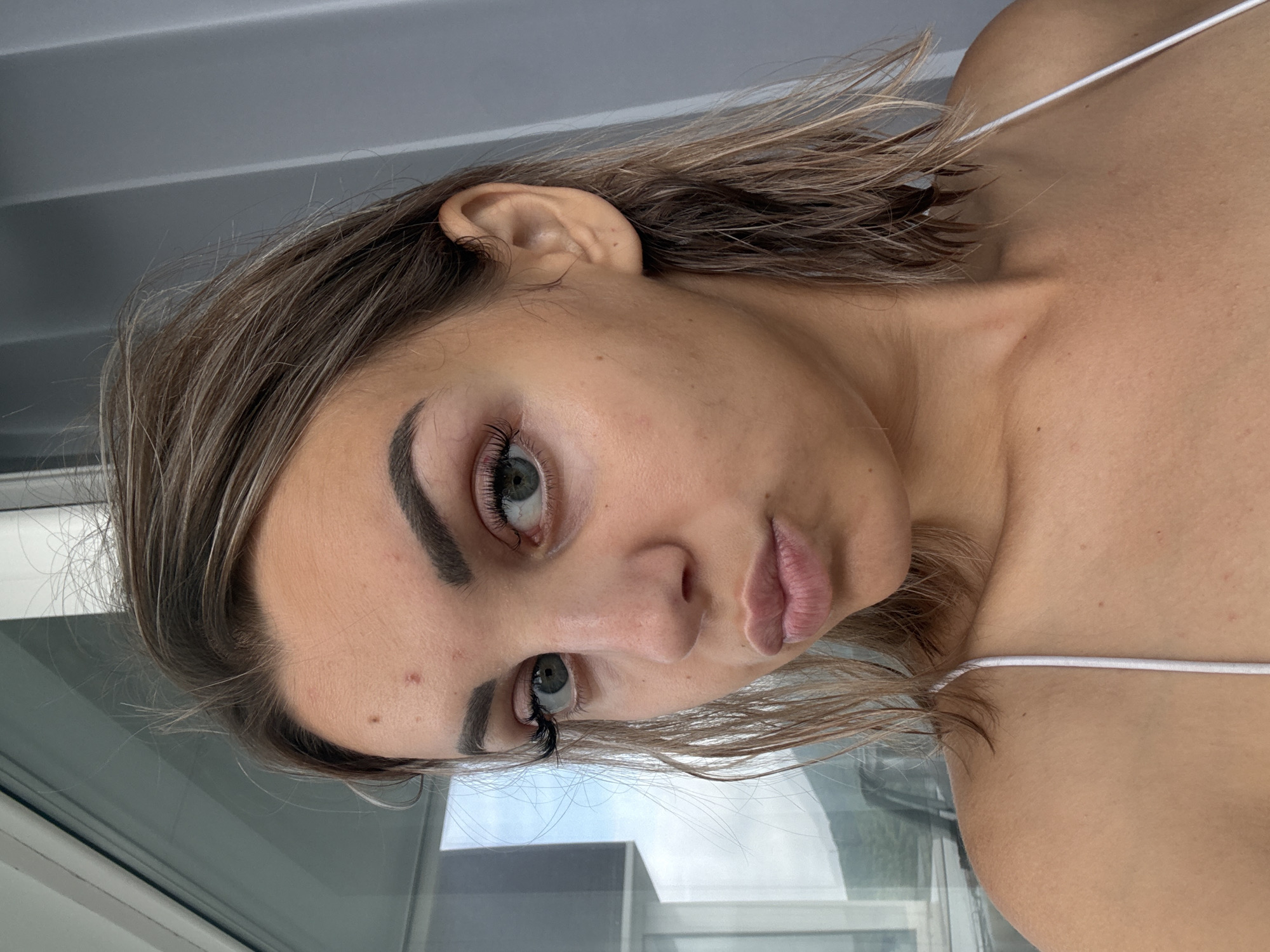 A young woman is captured in a close-up, head-and-shoulders shot, facing the camera with a slightly puckered expression. She has fair skin with some visible blemishes and pores on her forehead and cheeks. Her eyebrows are well-defined and dark, framing her light green eyes. She is wearing false eyelashes, which add a dramatic effect to her gaze. Her hair is light brown and appears damp, styled in loose, tousled waves that fall around her face and shoulders. She is wearing a thin, white spaghetti strap top, with one strap visible against her shoulder. The background consists of a blurry, neutral gray wall on the right and a window with a view of greenery and a body of water on the left. The lighting suggests an indoor setting, possibly a room with a large window. There is no discernible evidence of Kholmsk, Russia, or any other specific city in the image.
