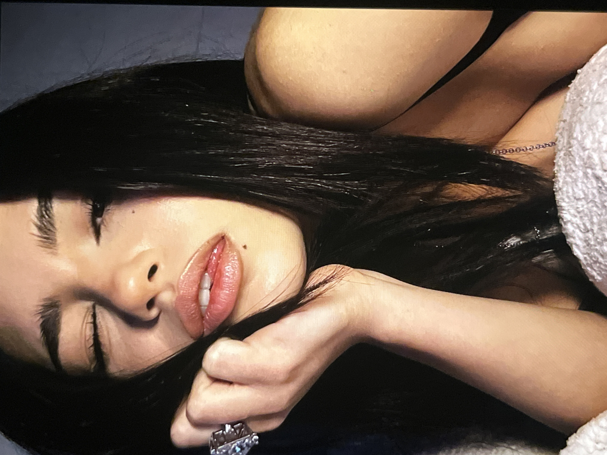 A close-up, head-and-shoulders shot of a young woman with her eyes closed and her lips parted. She has long, dark hair that partially obscures her face. Her right hand, adorned with a large, multi-faceted ring, is raised to her cheek. The lighting is soft and casts shadows, creating a moody atmosphere. The background is dark and indistinct, suggesting an indoor setting.