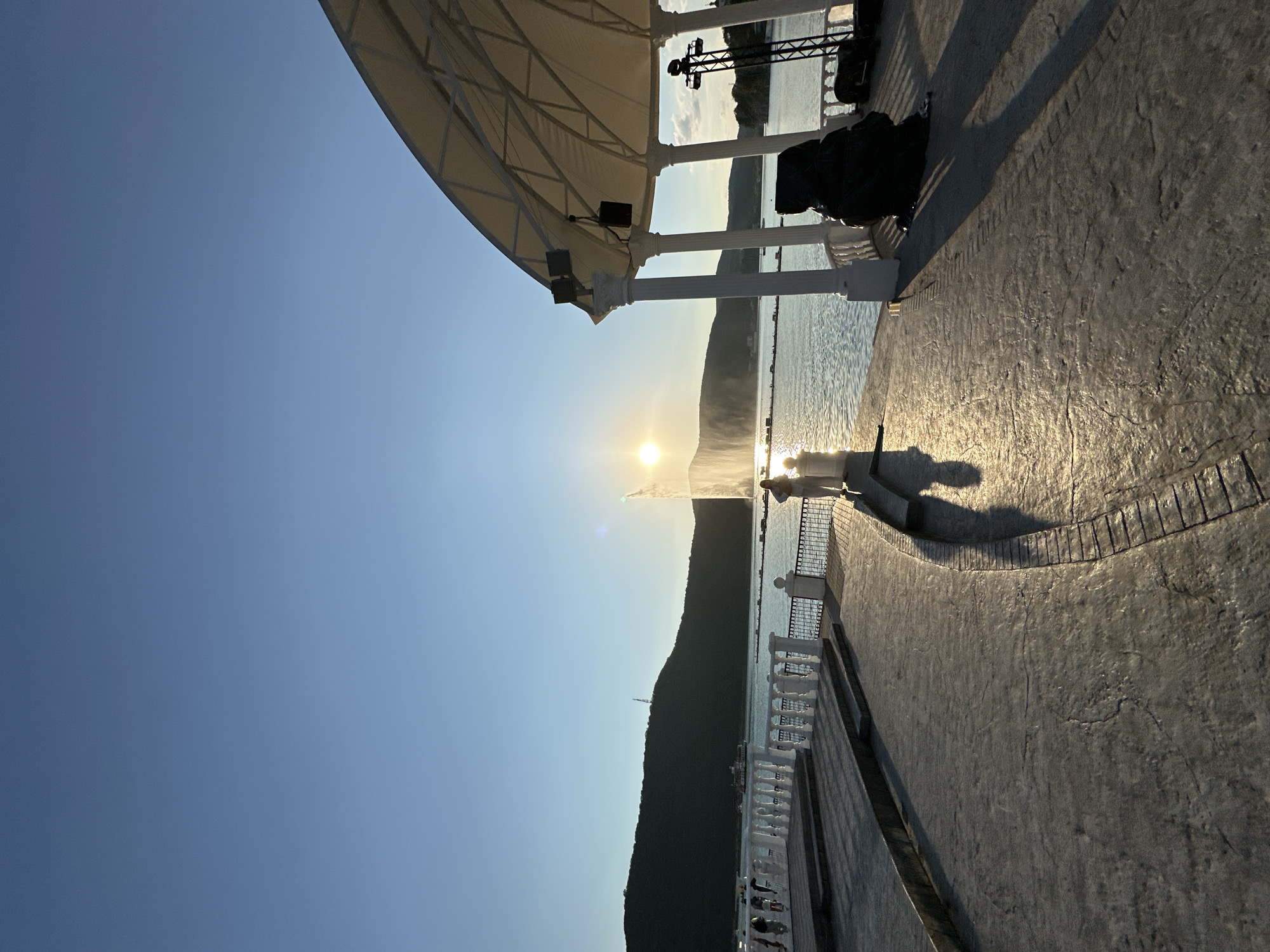 The image depicts a waterfront promenade at what appears to be sunset. The sun is low on the horizon, casting a warm glow and long shadows across the textured pavement. In the foreground, the promenade is lined with decorative white railings and columns, suggesting a developed tourist area. The sea stretches out to a dark, mountainous coastline in the background, partially silhouetted against the bright sky.
A large, white canopy structure with metal supports is visible on the left, providing shade. The water is calm, reflecting the sunlight, and a faint mist or spray rises from the surface, possibly from a fountain or sea spray. A few people are visible, mostly as silhouettes, standing and walking along the promenade, enjoying the view and the evening light.
On the far right side of the image, part of a stage or performance area can be seen, with lights and equipment, hinting at potential events or entertainment. The overall atmosphere is serene and picturesque, with the warm light of sunset creating a tranquil mood. The location is Abrau-Dyurso, Russia, known for its wineries and scenic coastline. The clear sky indicates fair weather.