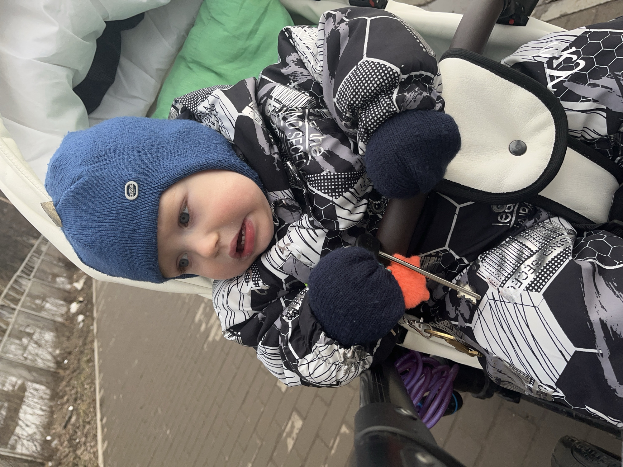 The image captures a young child seated in a stroller outdoors on a paved pathway in Chaikovsky, Russia. The child, an infant or toddler, wears a blue knitted beanie with a "CLASSIC" emblem, a black, white, and grey patterned winter jacket with geometric and textual designs, and dark blue knitted mittens. The child's mouth is slightly open, and their gaze is directed towards the upper right of the frame. Attached to the stroller's safety bar in front of the child are a set of keys with an orange fluffy keychain and a partially visible purple coiled cable lock. The stroller has a cream-colored interior lining, a green head cushion, and a cream-colored faux leather pouch with black trim and a central black snap button on its bar. The immediate foreground shows a paved path made of rectangular pavers. Beyond the path, a strip of bare ground with sparse, dry vegetation and remnants of snow is visible, backed by a light-colored metal fence structure and distant trees.