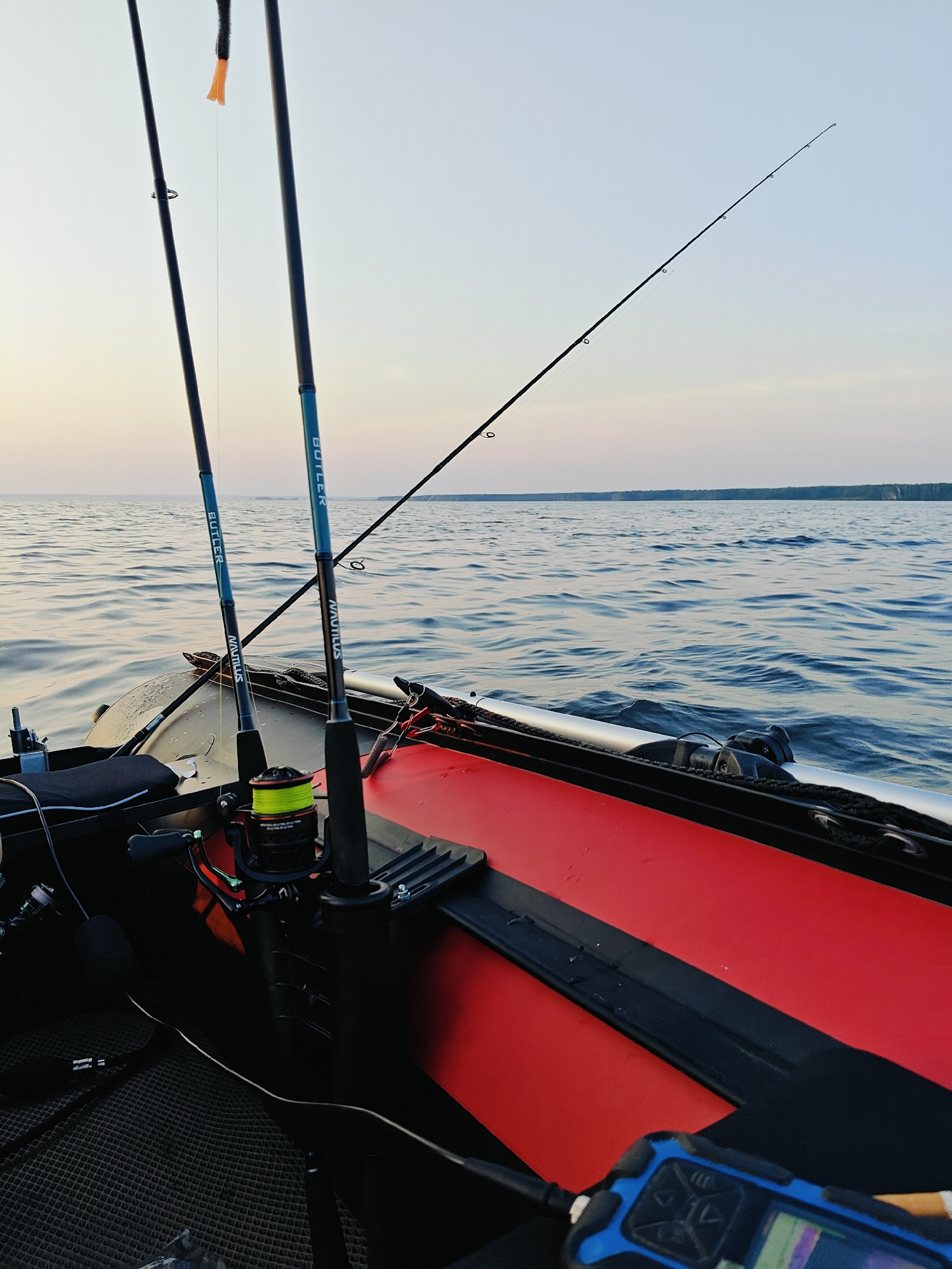 In the city of Chaykovsky, Russia, a view from an inflatable boat on a body of water shows fishing equipment. Two fishing rods are propped up in rod holders on the boat's deck. The rods have "BUTLER" and "NAUTILUS" branding. A fishing reel with bright yellow line is visible on one of the rods. The water is calm with small ripples, and the sky is clear and pale. The edge of the red inflatable boat and black seating are also in the frame. A small, blue electronic device with a screen and buttons is in the foreground. The horizon line suggests a calm day with distant landmass.