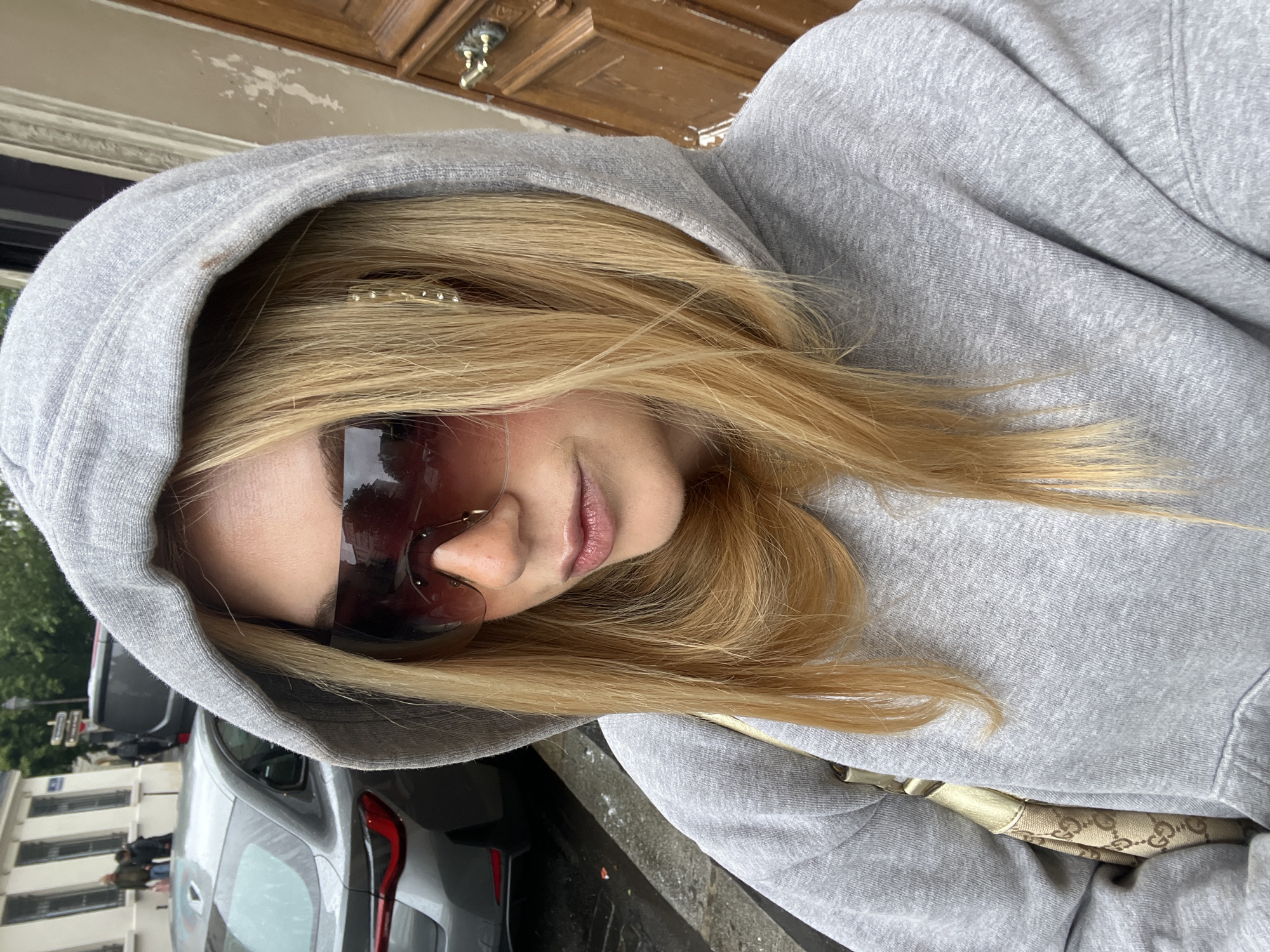 A woman with blonde hair wearing sunglasses and a gray hoodie is positioned in front of a parked gray car. The background reveals a building with multiple windows, and across the street, a tree is visible. The location is inferred to be in Paris, France based on the architectural style of the building. A wooden door is visible in the upper portion of the image. The woman is holding a patterned bag.