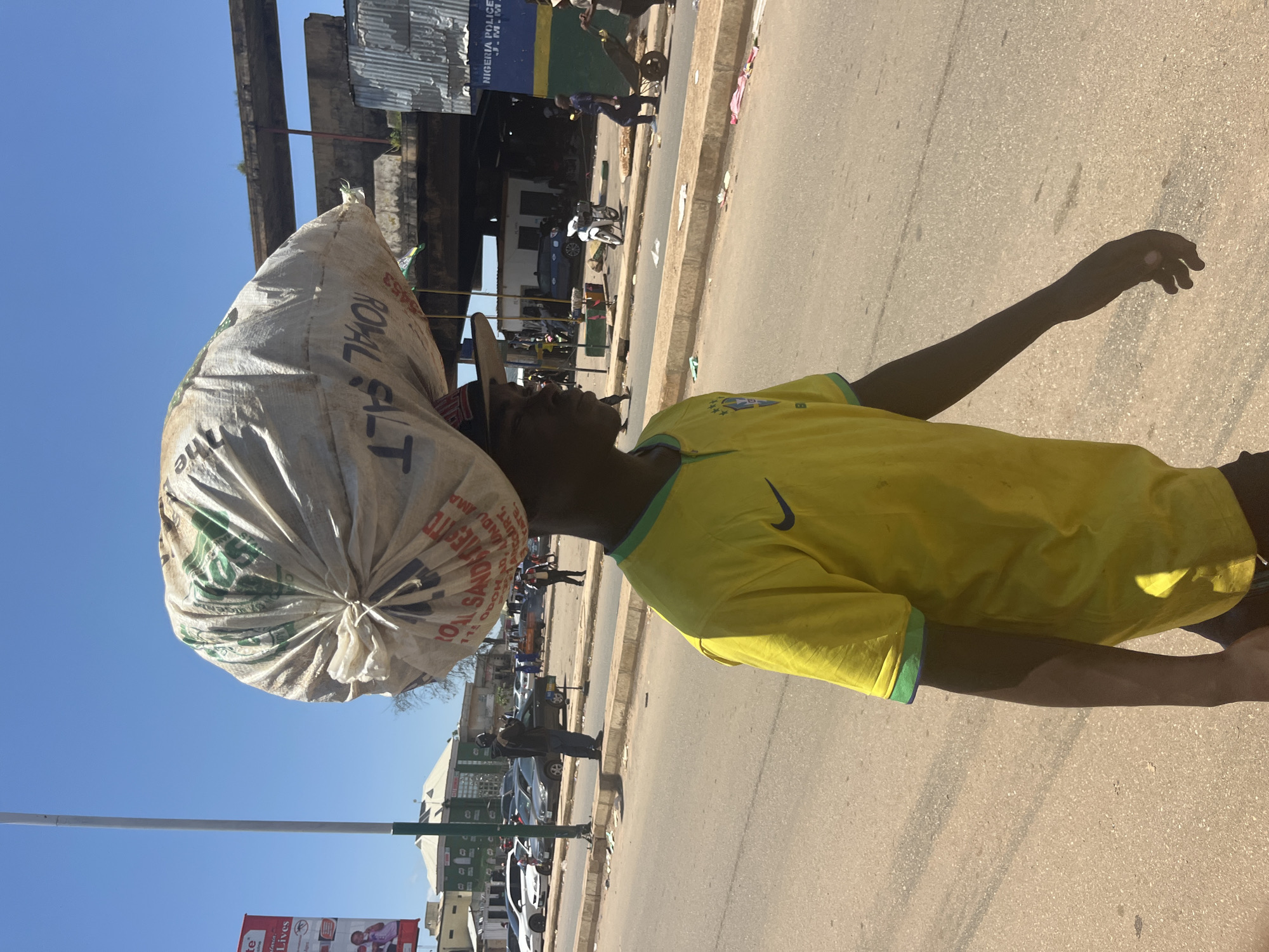 In Jos, Nigeria, a man walks along a road carrying a large, white bag of salt on his head. The bag has green lettering that reads "ROYAL SAND...SALT". He is wearing a yellow t-shirt with a green and blue collar, and a dark baseball cap. The road appears to be a main thoroughfare with parked cars and other pedestrians in the background. A sign in the distance reads "NIGERIA POLICE". The sky is clear and blue, indicating a sunny day. The scene depicts everyday life in an urban setting in Nigeria.