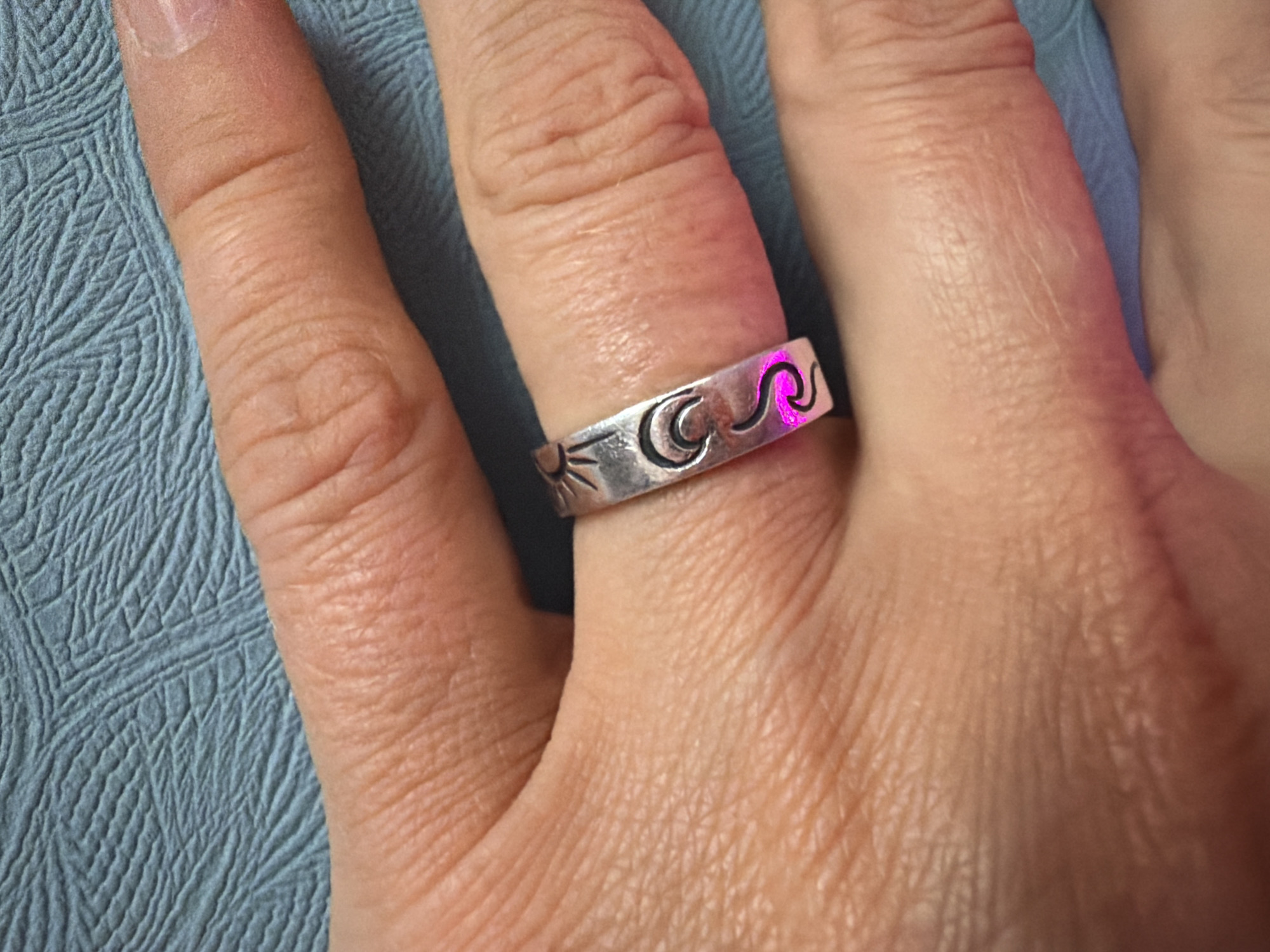 A close-up, high-angle photograph focuses on an adult's left hand, resting on a textured light blue surface. The hand's skin, a light to medium tone, shows natural lines and creases across the fingers and knuckles. The fingernail of the index finger is visible, appearing natural or with a clear polish.
A notable silver-toned band ring is worn on the middle finger. The ring is wide and features a sequence of engraved or embossed symbols: a stylized sun on the far left, followed by a crescent moon, and then two distinct wavy lines. A bright magenta or pink light prominently illuminates the wavy lines on the ring, suggesting a specific lighting effect or reflection.
The background consists of a soft, light blue material with an intricate, geometric textured pattern, possibly a fabric or mat. The setting appears to be indoors, under artificial lighting, with no specific time of day discernible. No particular activity is captured beyond the display of the hand and ring. No text is visible in the image. This scene is contextualized as being in Saint Petersburg, Russia.