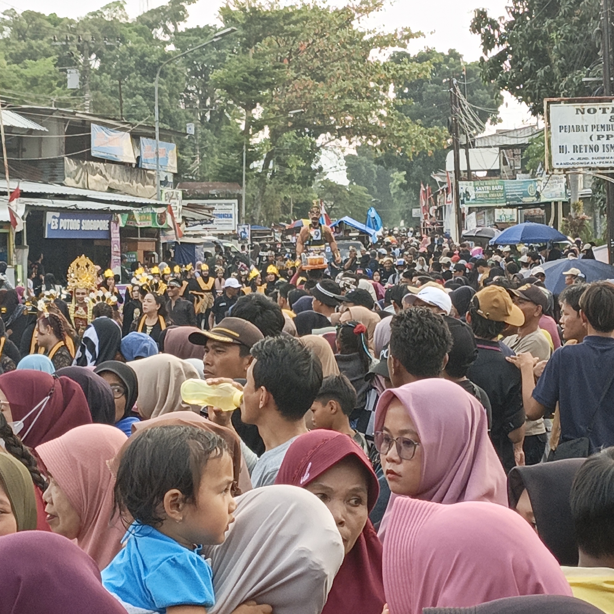 A crowded street scene is depicted in Randudongkal, Indonesia, likely during a festival or parade, judging by the celebratory attire and the large gathering of people. In the foreground, several individuals are visible, including a young child in a blue shirt, an adult woman wearing a maroon hijab and glasses, and another woman in a pink hijab. Their gazes are directed off-frame, suggesting they are observing something happening in front of them.
The middle ground is filled with a dense crowd of people, many wearing dark clothing, some with yellow accents or hats, and others in traditional attire. A central figure stands out, adorned in a golden headdress and ornate clothing, possibly a performer or a decorated individual. They are holding a tray with beverages. Another individual dressed similarly with a golden headdress is positioned behind them. Several blue umbrellas are visible above the crowd, indicating possible sun protection or the beginning of rain. Banners and signs are displayed along the buildings lining the street, with some text visible, including "ES POTONG SINGAPORE" and what appears to be a sign for "PJABAT PEMBU" which could be "Pejabat Pembantu" or similar.
The background features a street lined with modest buildings and vegetation, suggesting a semi-urban or town setting. Power lines crisscross overhead, supported by utility poles. The overall atmosphere appears lively and communal.