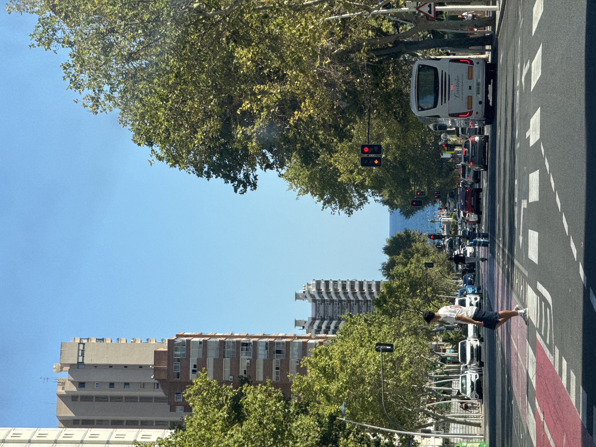 The image depicts a street scene in Benidorm, Spain, on a clear day. The view is down a wide avenue lined with tall apartment buildings and lush green trees. In the foreground, a pedestrian is crossing a busy street at a marked crosswalk. Several cars and a white tour bus are visible on the road, some stopped at a traffic light, indicating moderate traffic flow. The bus has the name "Castaño" and contact information on its rear. Further down the street, the blue of the Mediterranean Sea is visible in the distance. The architecture suggests a resort town, with the buildings characteristic of coastal Spanish cities. The sky is a bright, cloudless blue.