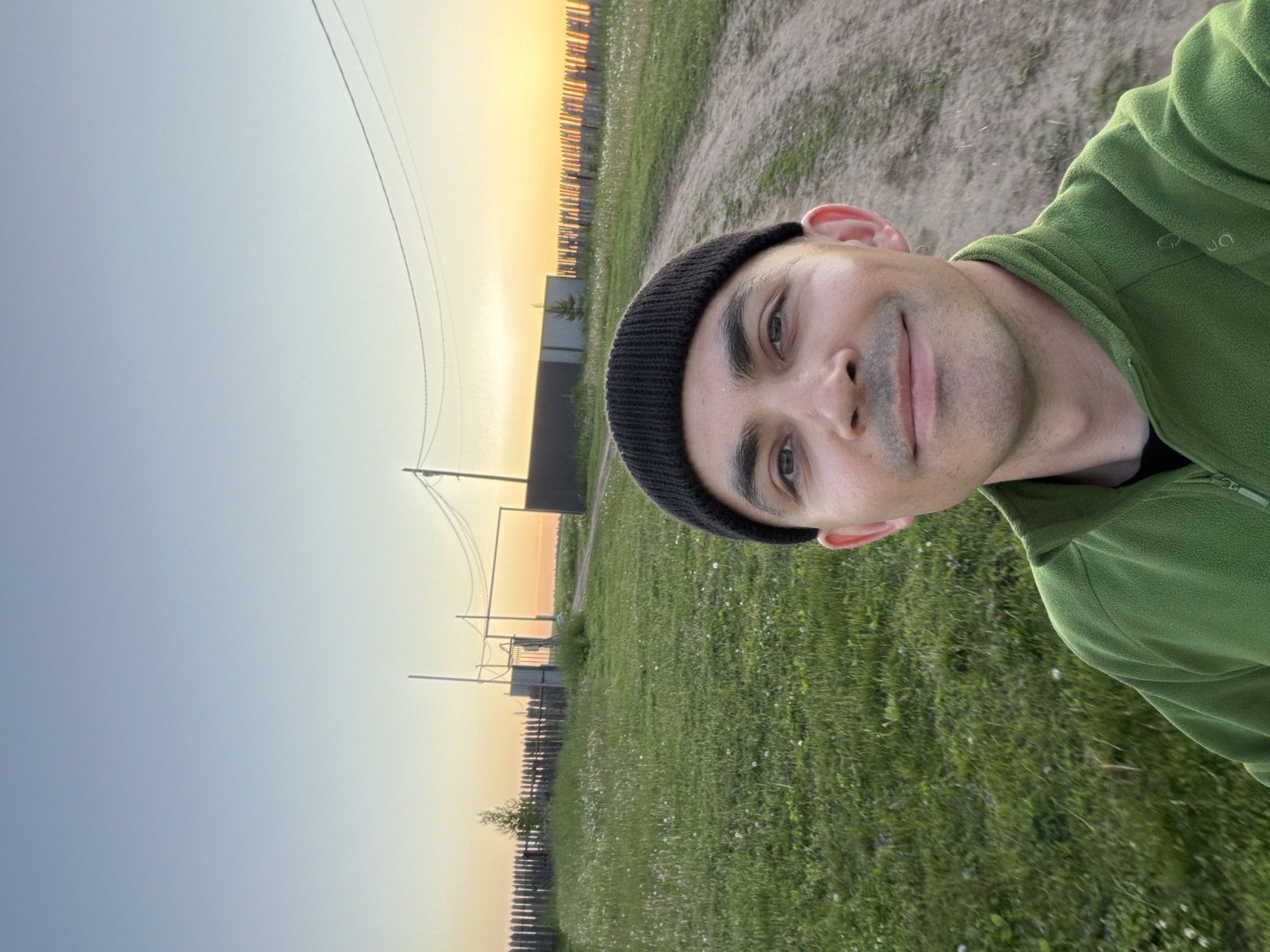 The image is a selfie taken by a man outdoors during the golden hour, likely sunset, in a rural area of Kechur, Russia. The man, with a slight smile and subtle stubble, looks directly at the camera. He is wearing a black knit beanie and a green fleece jacket, which features a white logo resembling "QUECHUA" on the right shoulder.
The scene behind him depicts a rustic landscape. To his left, a dirt path is visible. The foreground and midground are covered in green grass dotted with small white flowers. A low wooden picket fence stretches across the midground horizontally. Beyond the fence, utility poles with overhead power lines, some indistinct building structures, and a small tree can be seen.
The sky transitions from a pale blue-grey on the left to a warm, vibrant orange-yellow on the right, indicating the low angle of the sun. The overall impression is one of calm and tranquility in a natural, countryside setting.